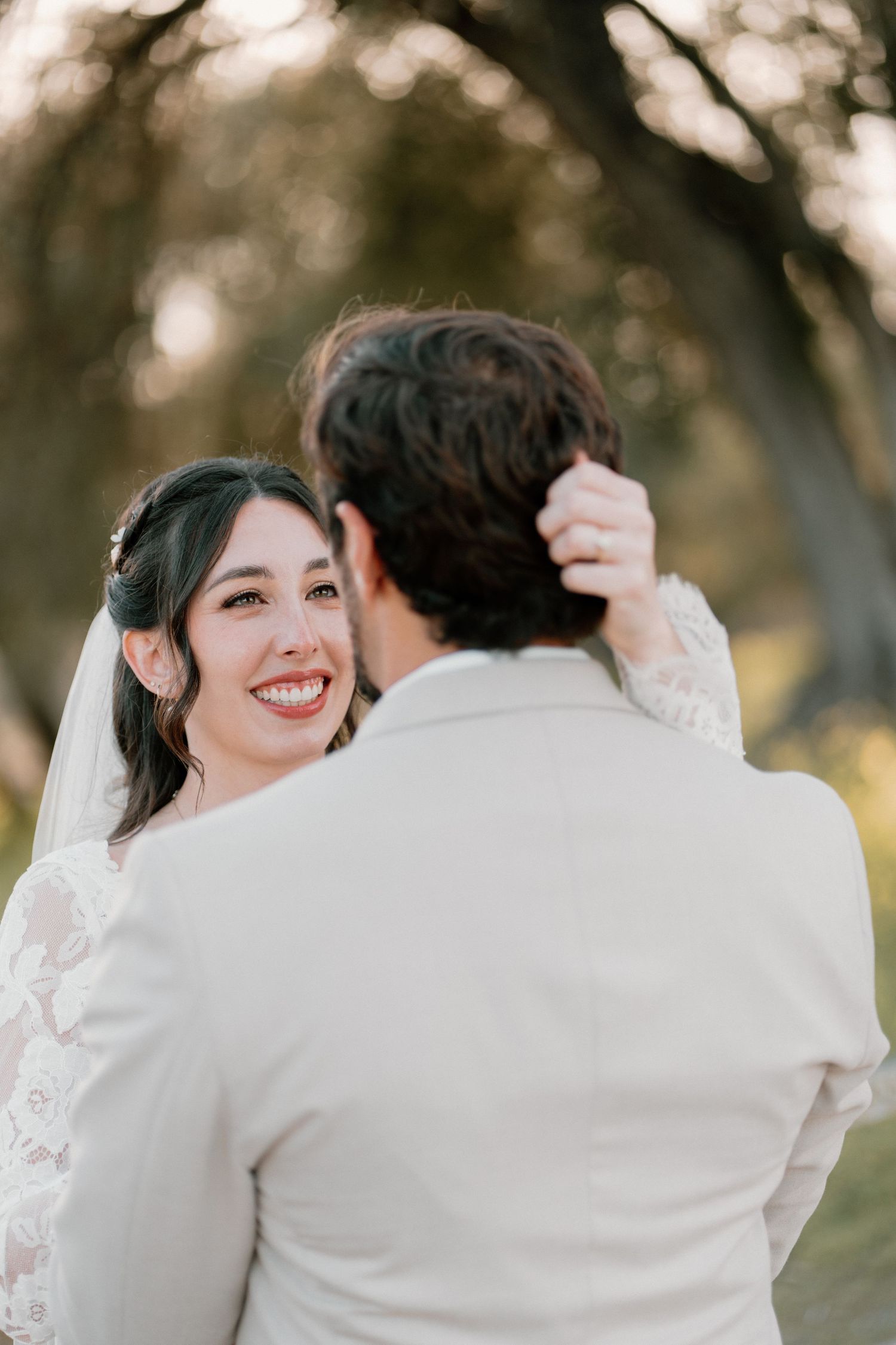 Smiling bride in lace wedding dress gazing lovingly at groom while touching his hair outdoors.