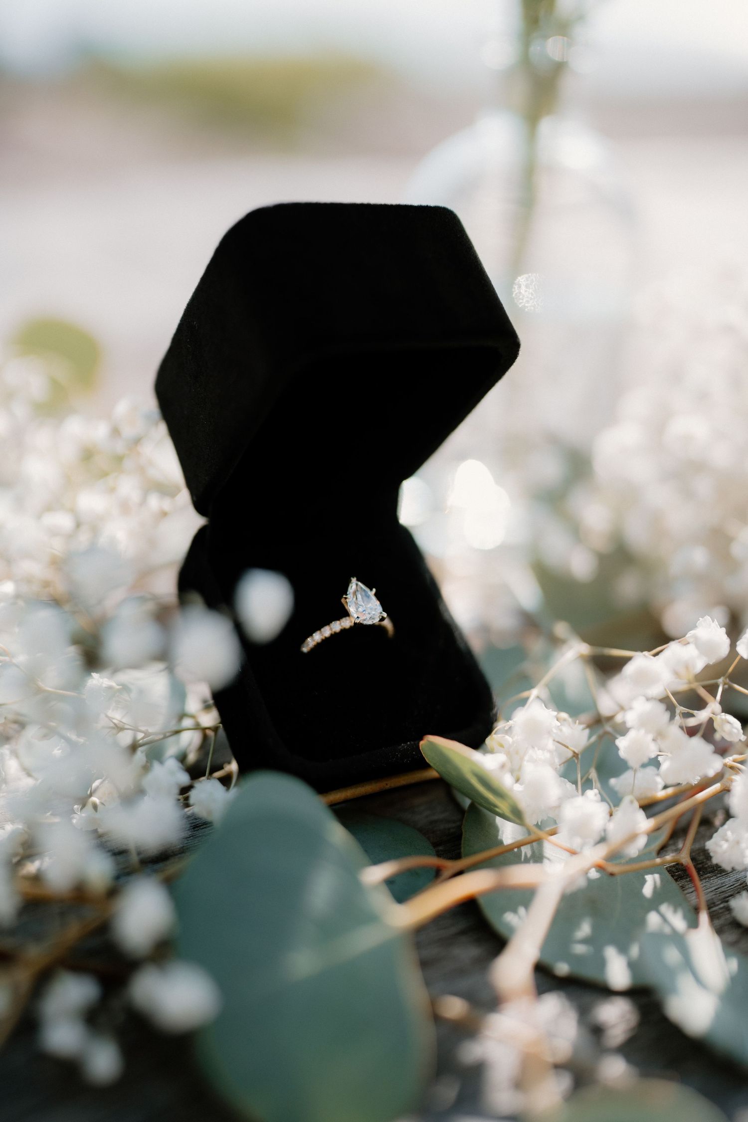 Diamond solitaire engagement ring in a black velvet box nestled among white baby's breath blooms.