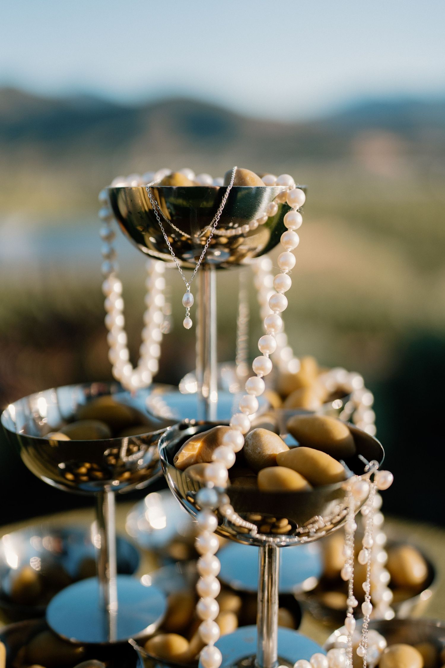 Elegant champagne tower coupe glasses filled with gold chocolates and draped with pearl necklaces outdoors.