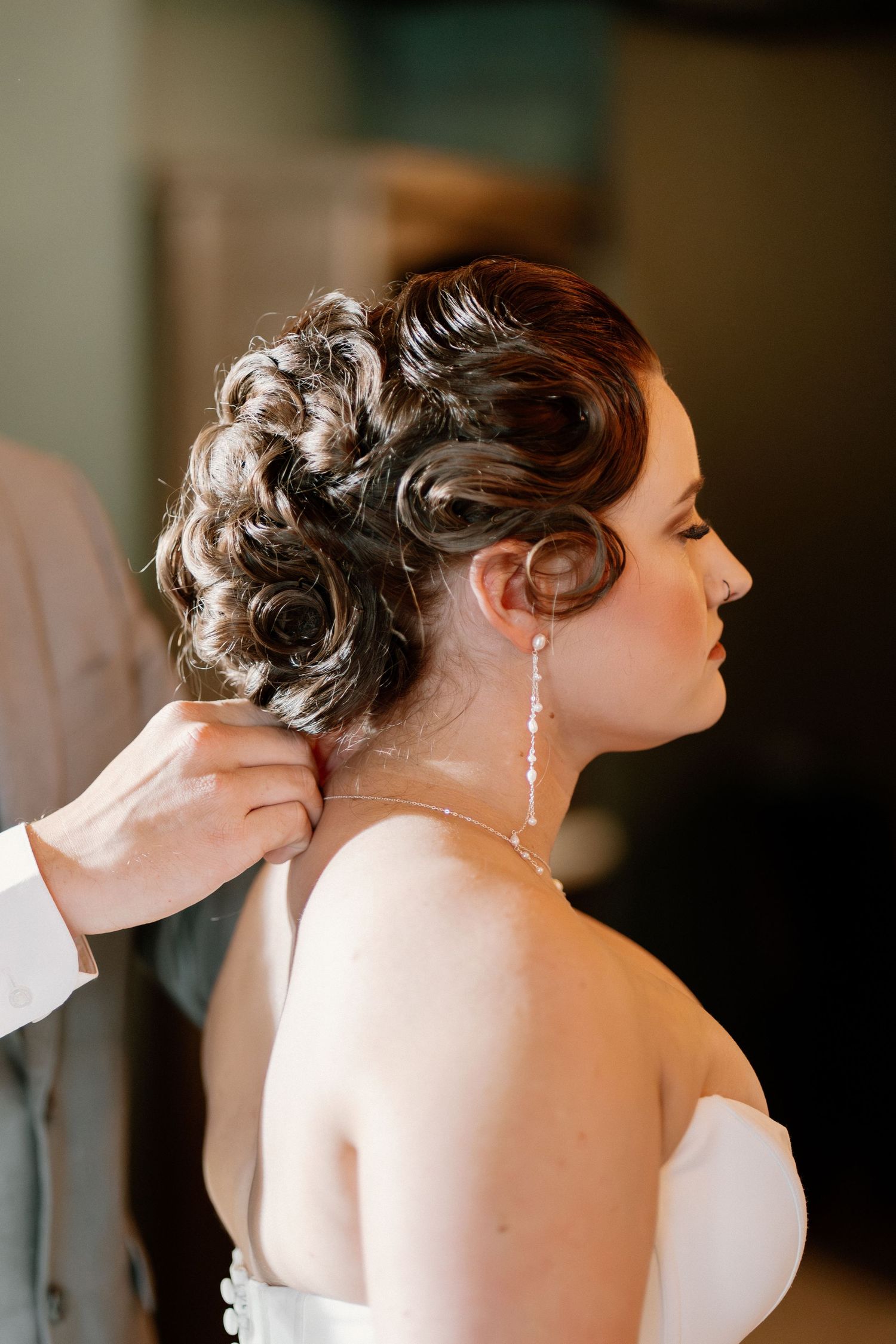 Bride with elegant curled updo hairstyle getting necklace fastened, wearing drop earrings and strapless white gown.