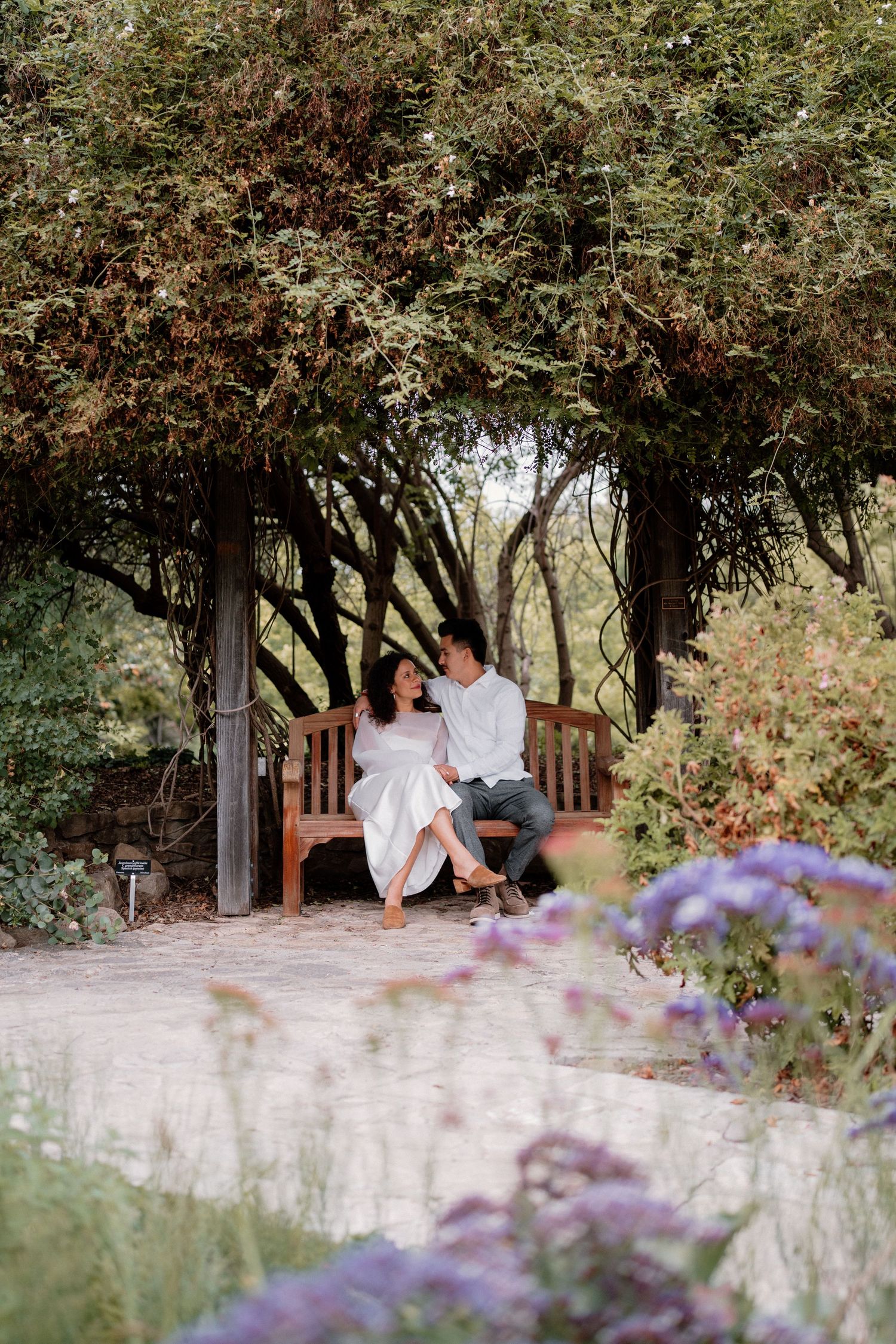 A couple sits together on a wooden bench nestled under trees in a beautiful garden setting.