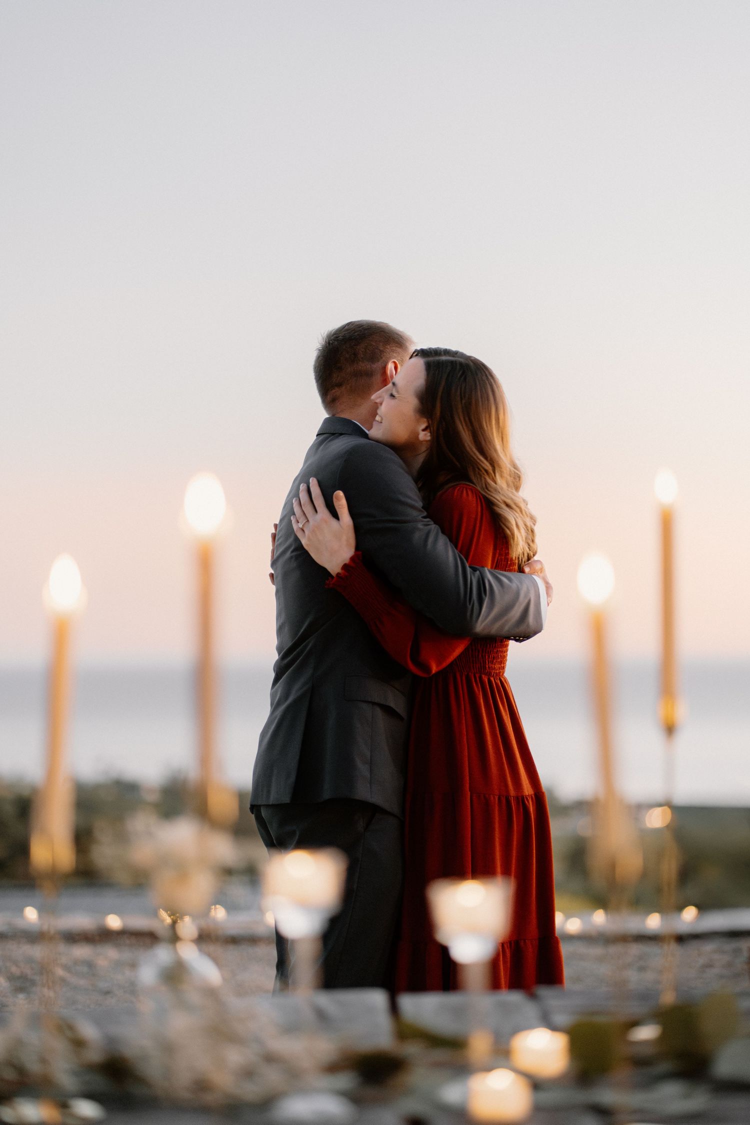 Couple sharing a romantic kiss surrounded by tall candles at an outdoor proposal setup by the sea.
