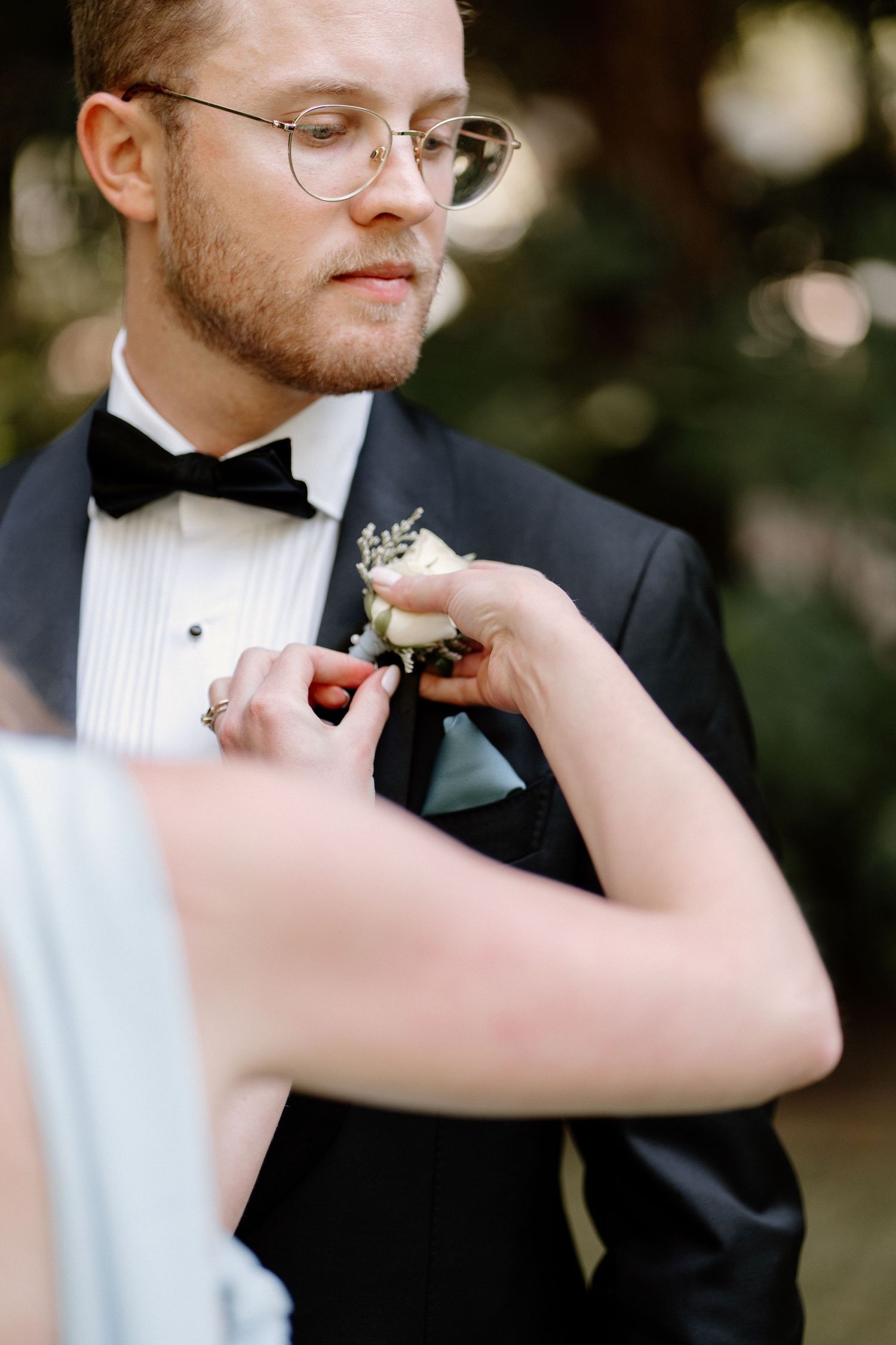 Bride pins a white rose boutonniere onto groom in black tuxedo with bow tie outdoors.