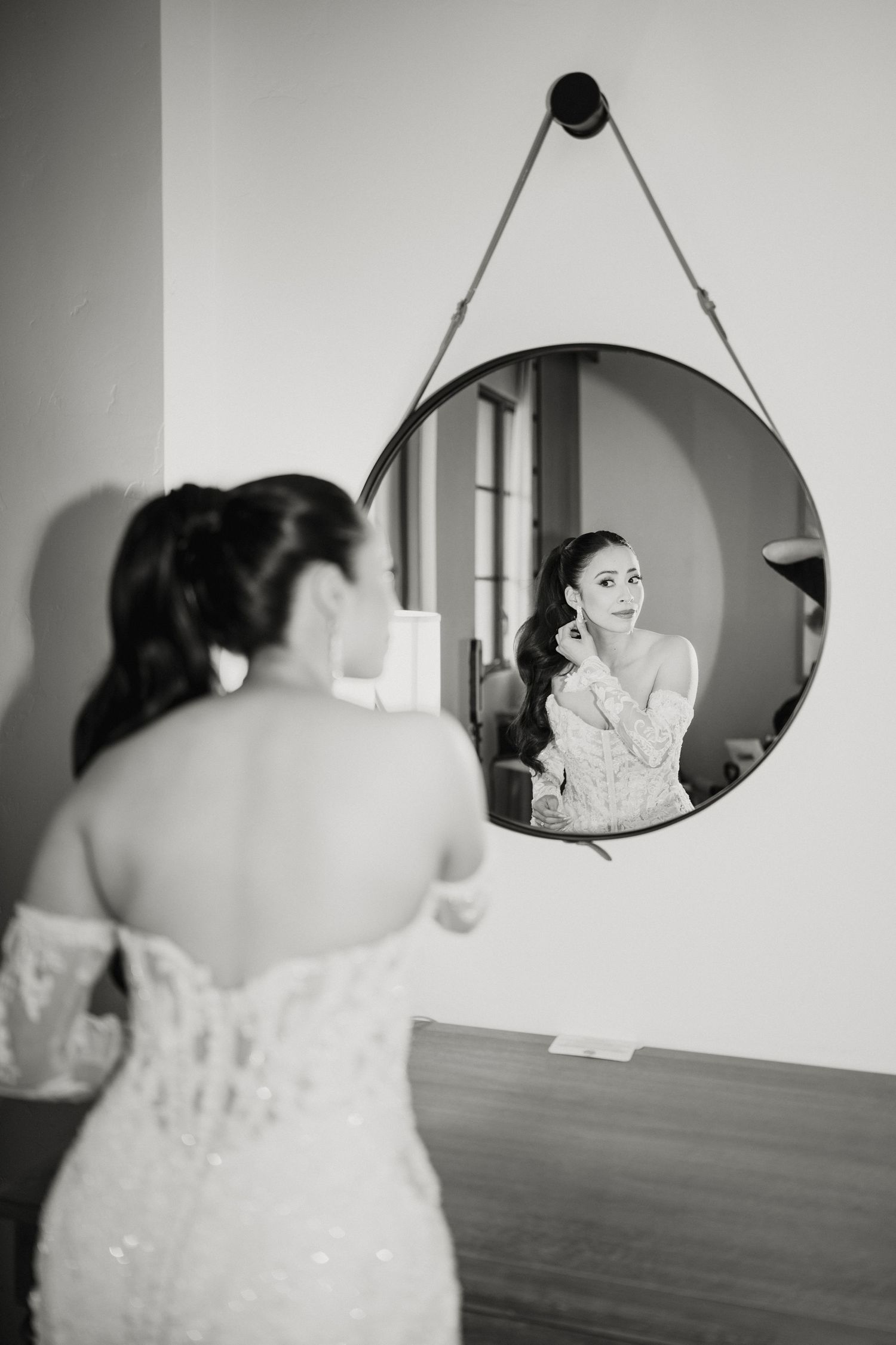Elegant bride in lace wedding gown adjusting earring while gazing into a round hanging mirror in black and white photo.