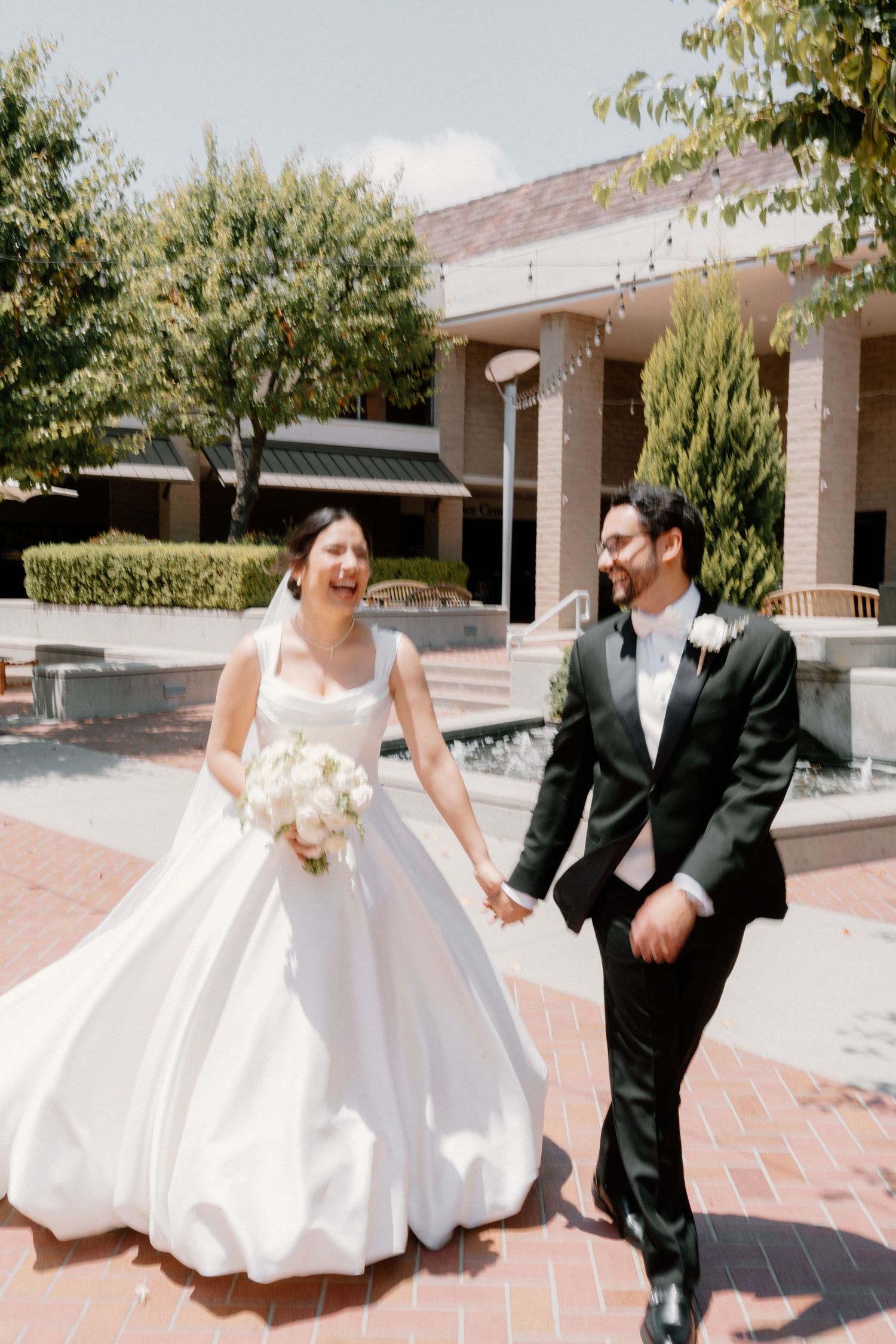 Happy bride and groom hold hands walking outdoors in classic white gown and black tuxedo on wedding day.