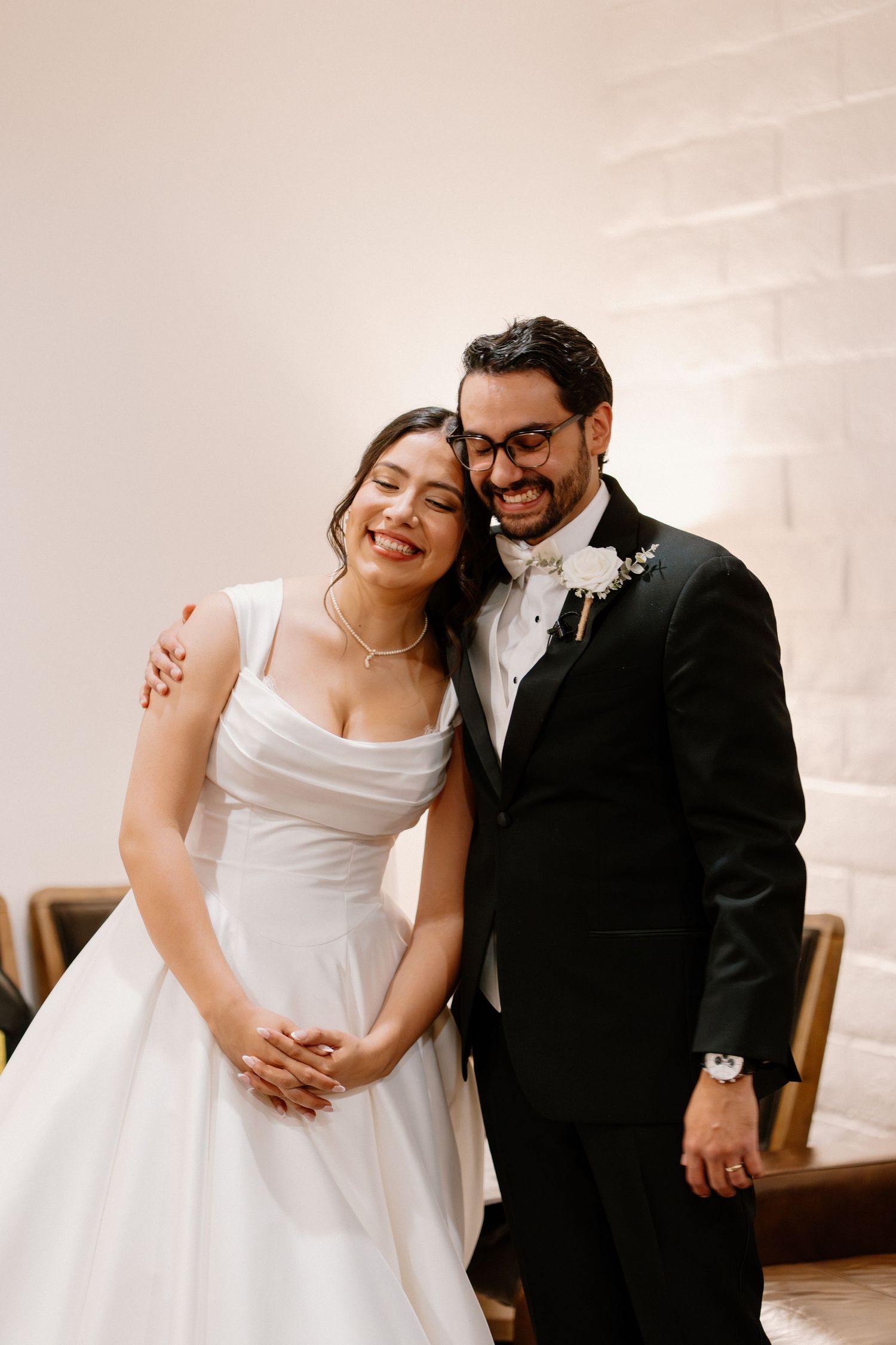 Smiling bride and groom pose together indoors, bride in white gown and groom in black tuxedo.