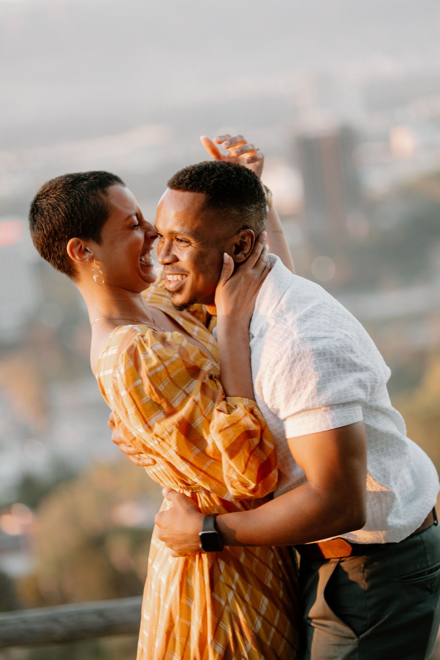 A joyful Black couple embraces and laughs together at golden hour with a city skyline in the background.
