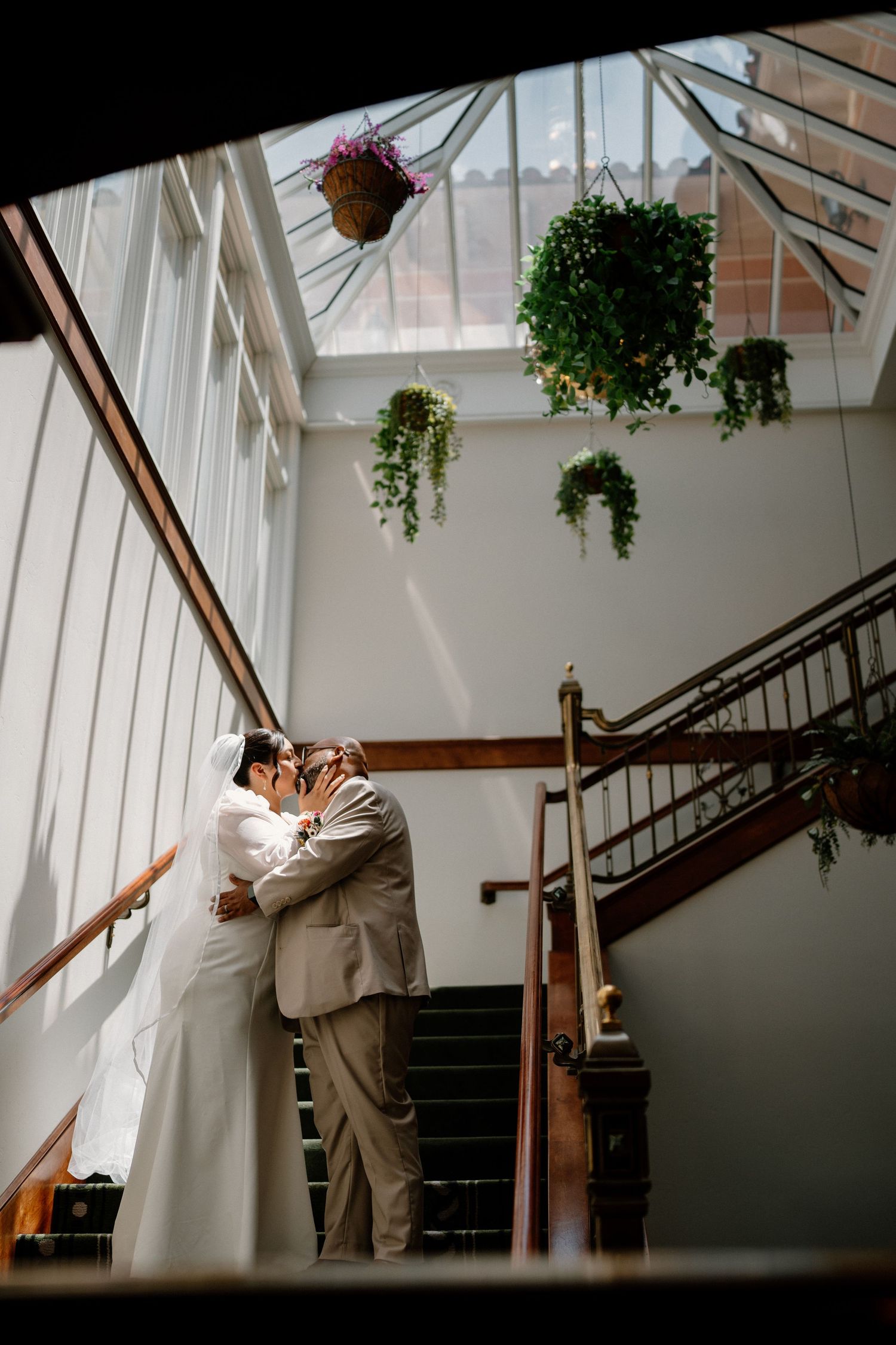 Bride and groom sharing a kiss on staircase under skylight with hanging plants.