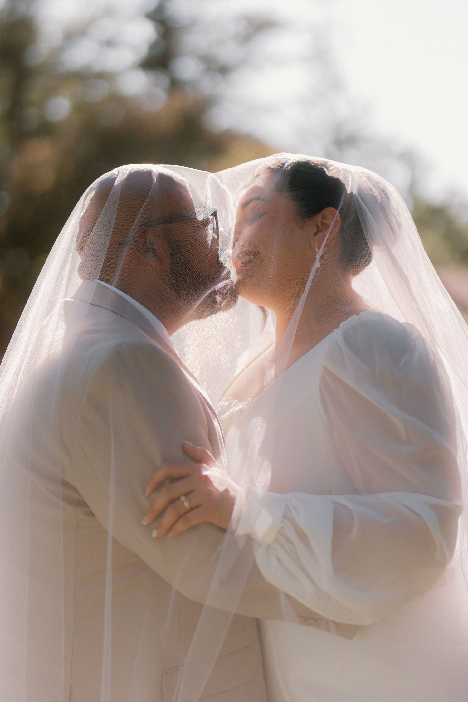 Bride and groom share a romantic kiss under a sheer wedding veil in soft natural light.