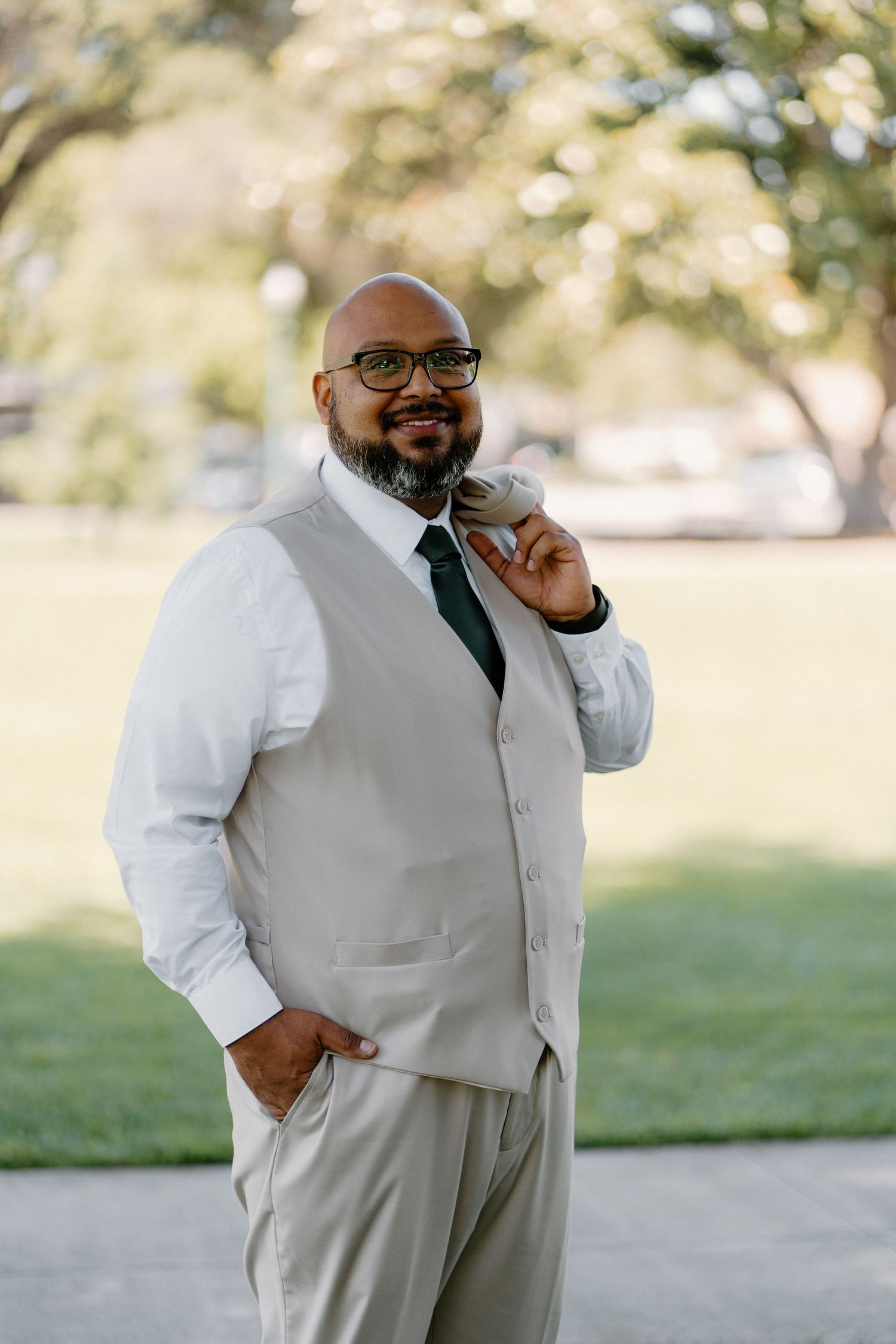Smiling bald man with beard and glasses wearing a light gray vest suit with dark tie outdoors.