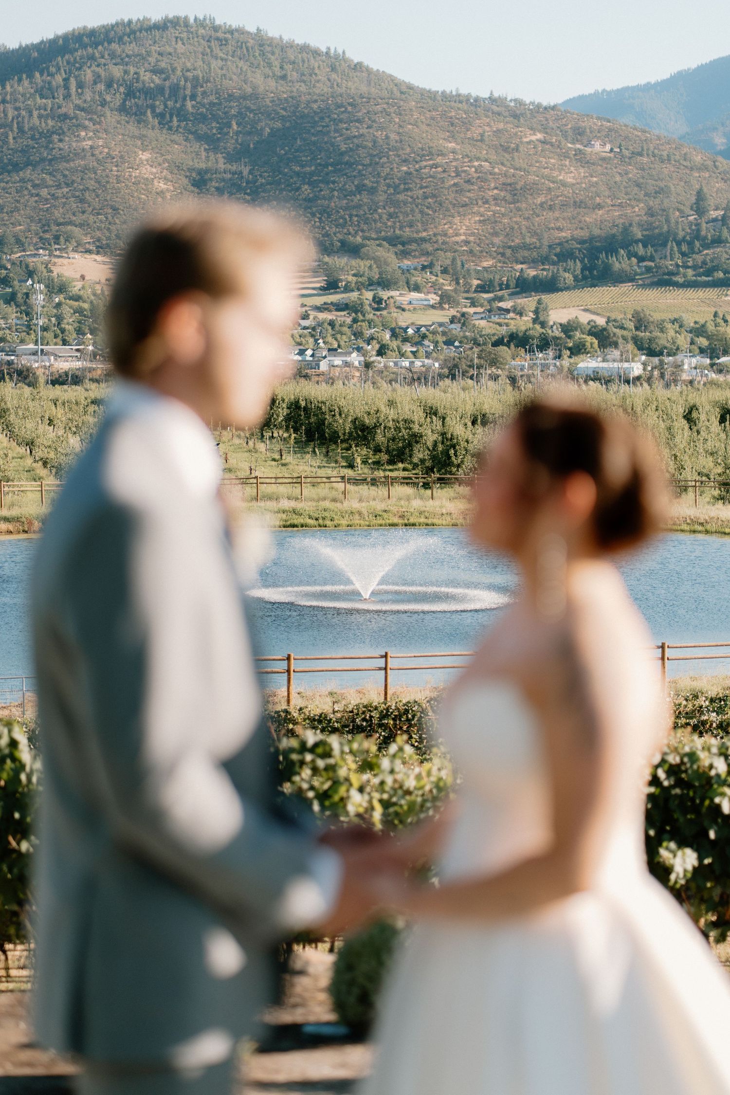 Bride and groom hold hands with a scenic lake fountain and mountain backdrop at an outdoor wedding venue.