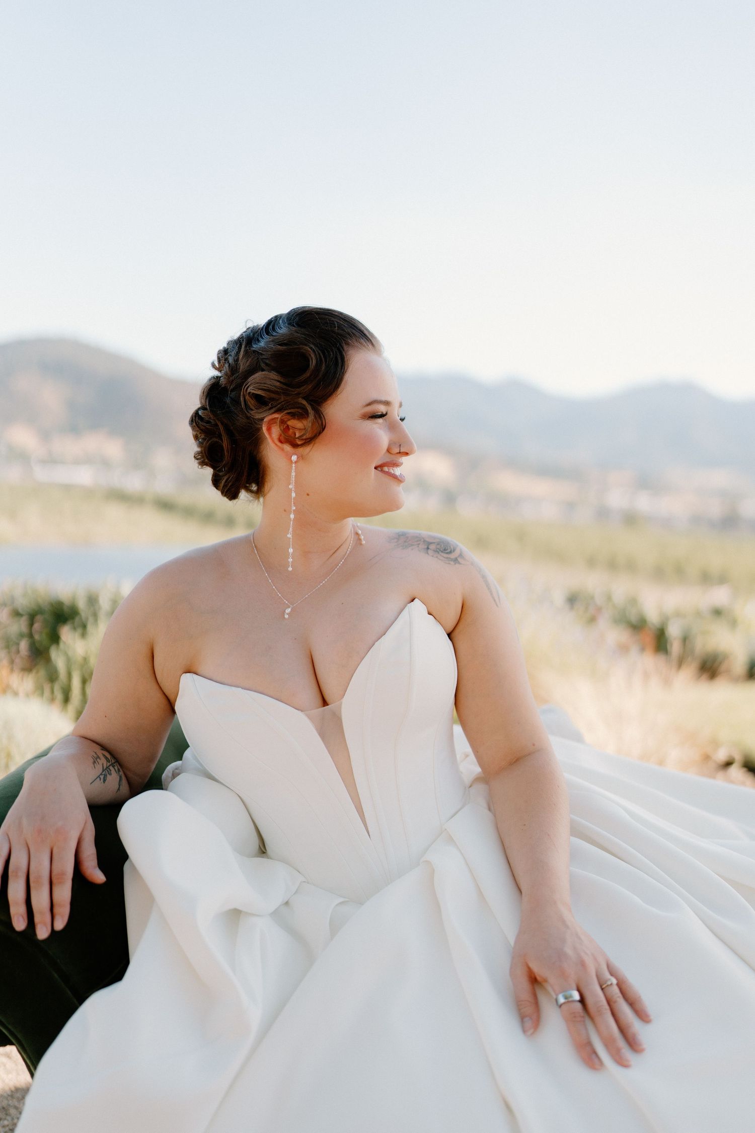 Smiling bride in strapless white gown with updo and dangling earrings against mountain view.