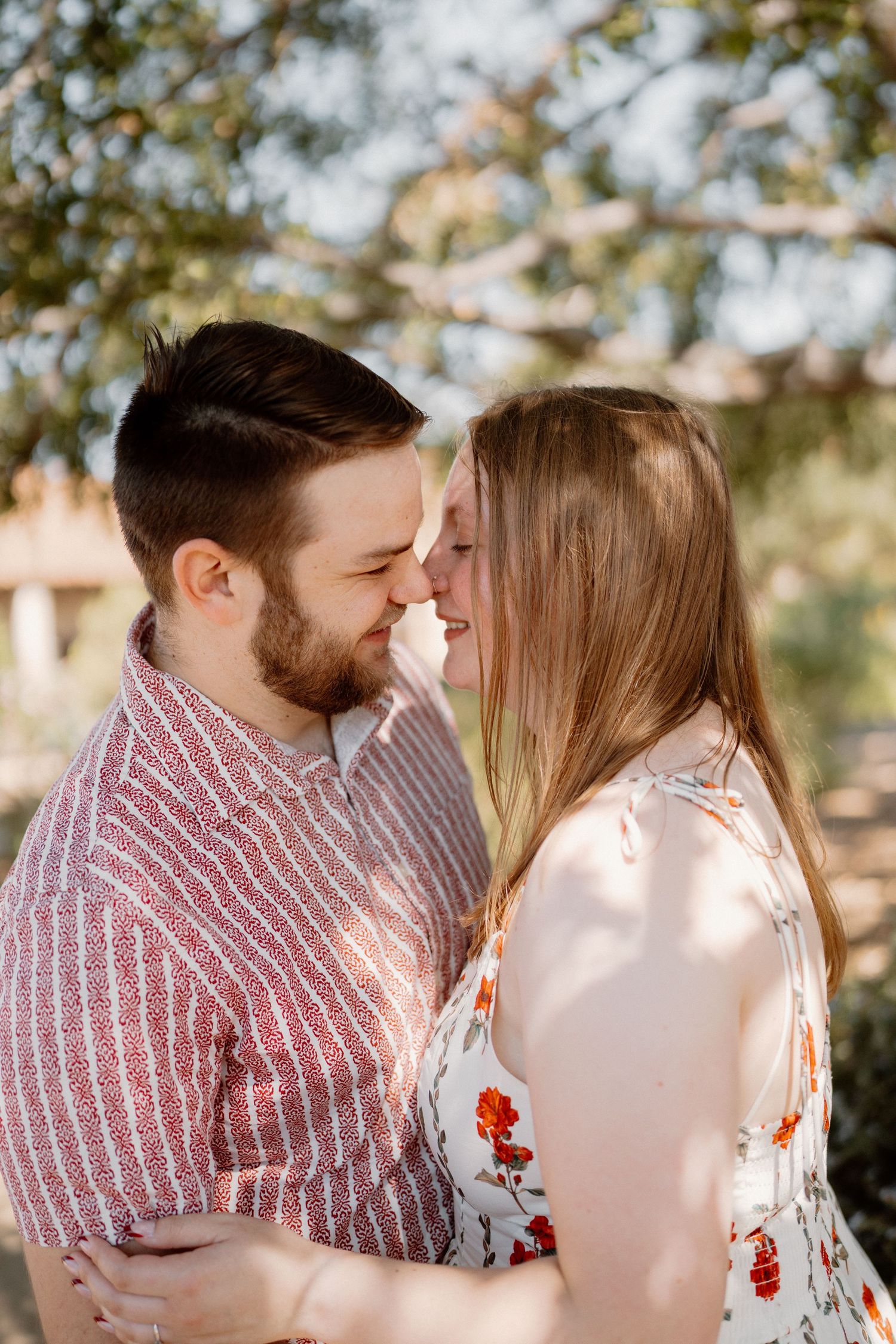 A couple shares an intimate nose-to-nose moment outdoors, embracing under dappled sunlight.