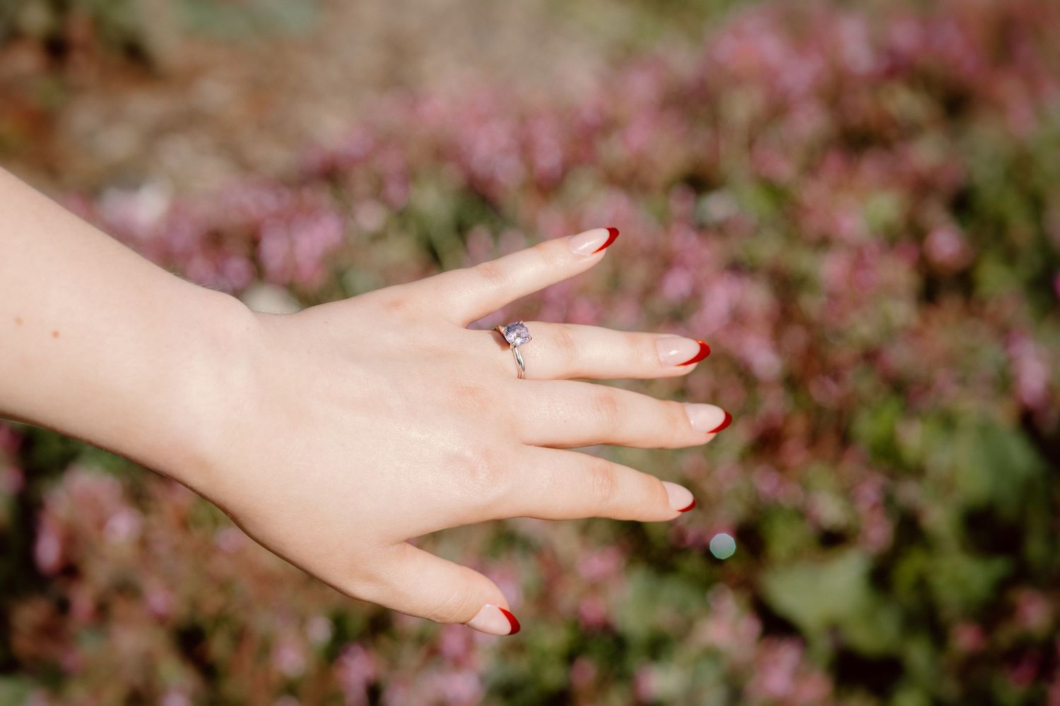 Close-up of a woman's hand with red nails wearing a diamond engagement ring against a floral background.