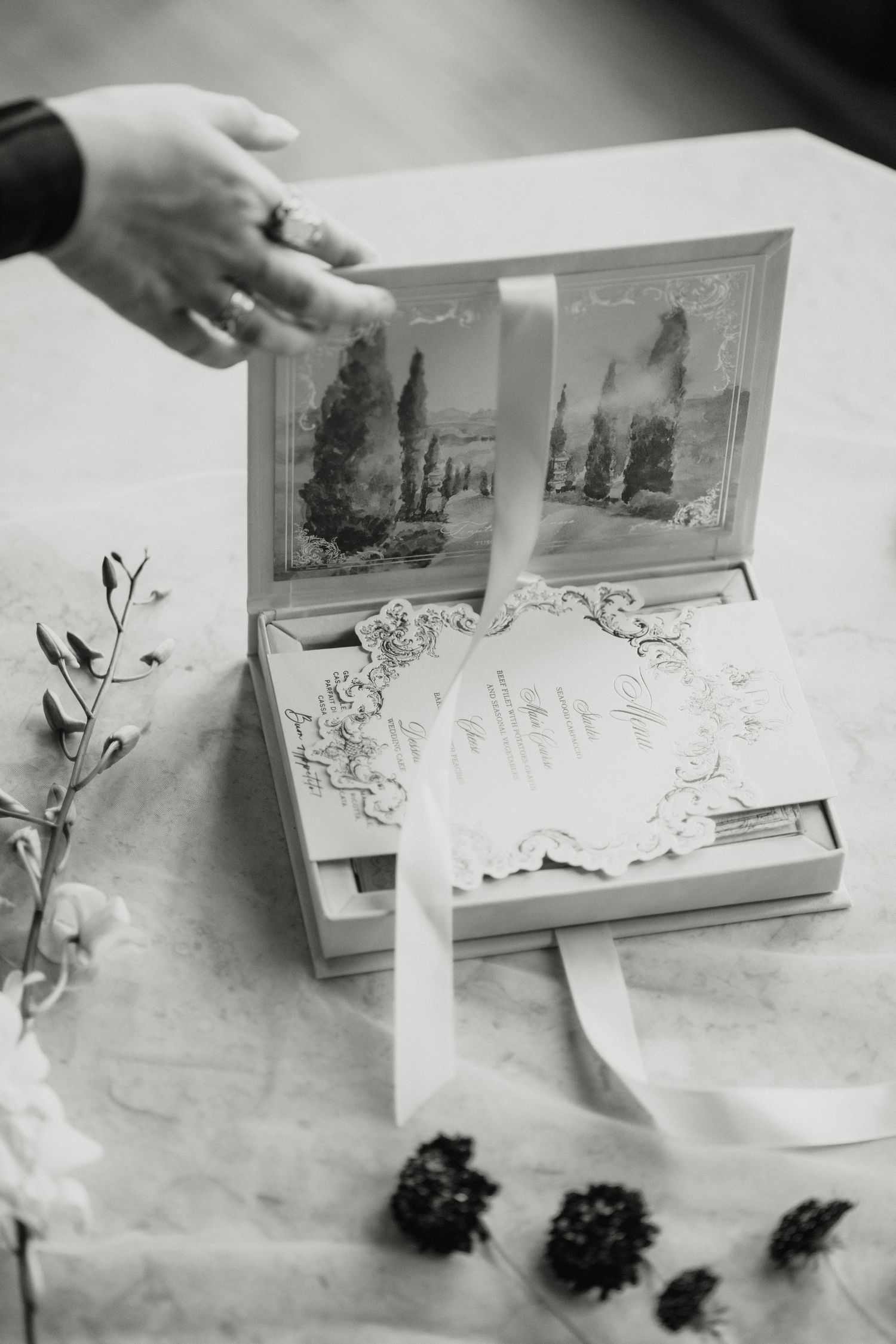 Black and white photo of elegant wedding invitation box with ribbon and floral details on marble surface.