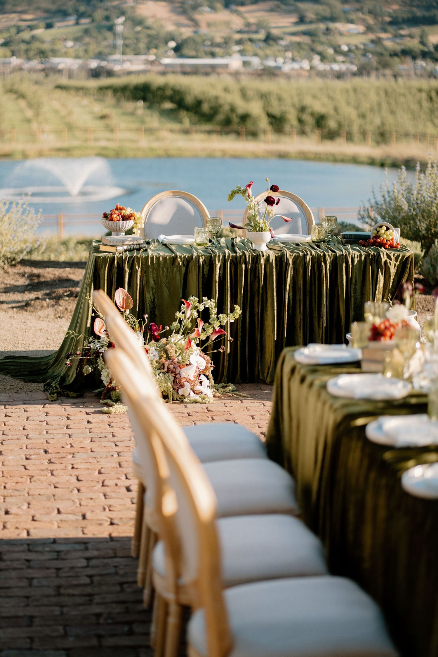 Outdoor wedding sweetheart table with olive velvet draping, floral arrangements, and scenic lake view.