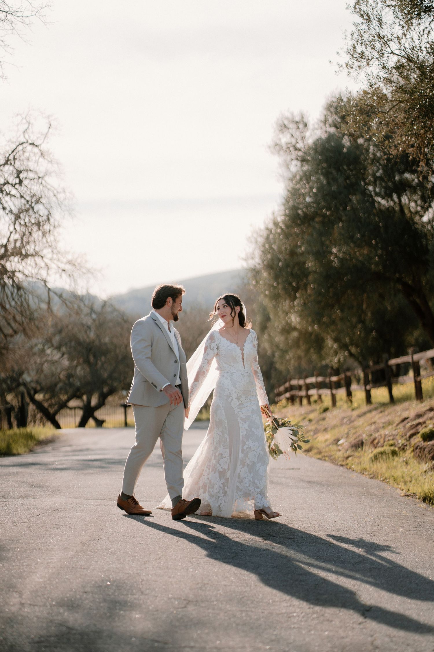 Bride in lace gown and groom in grey suit walking together on a scenic outdoor road on their wedding day.