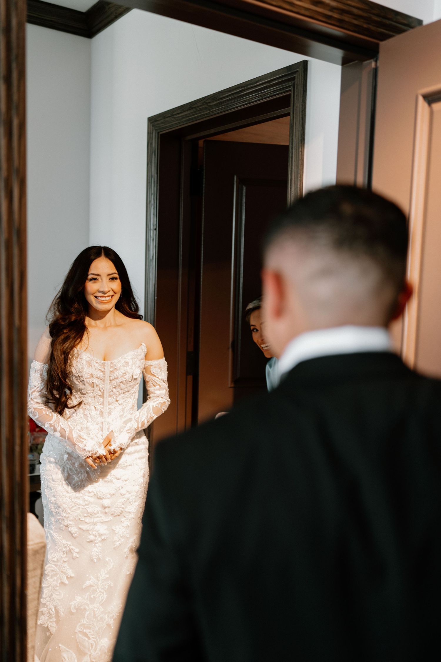 Bride in lace wedding gown smiling at groom during emotional first look moment indoors.