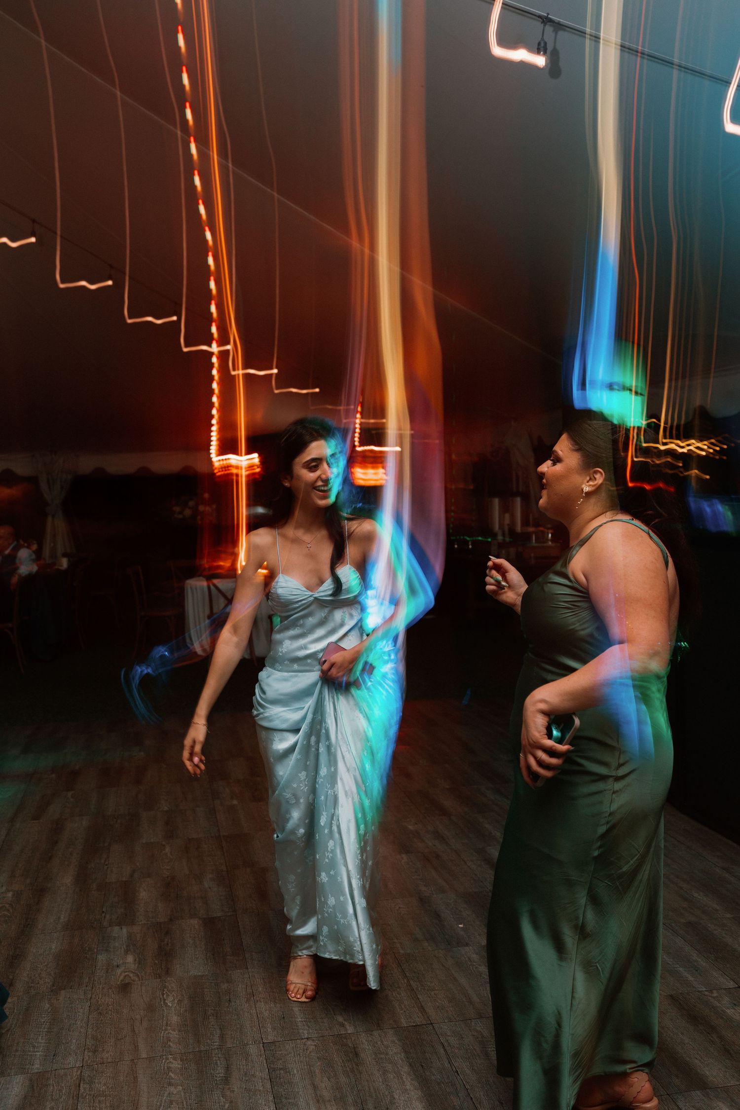 Two women in elegant gowns dancing with colorful light streaks at a wedding reception.
