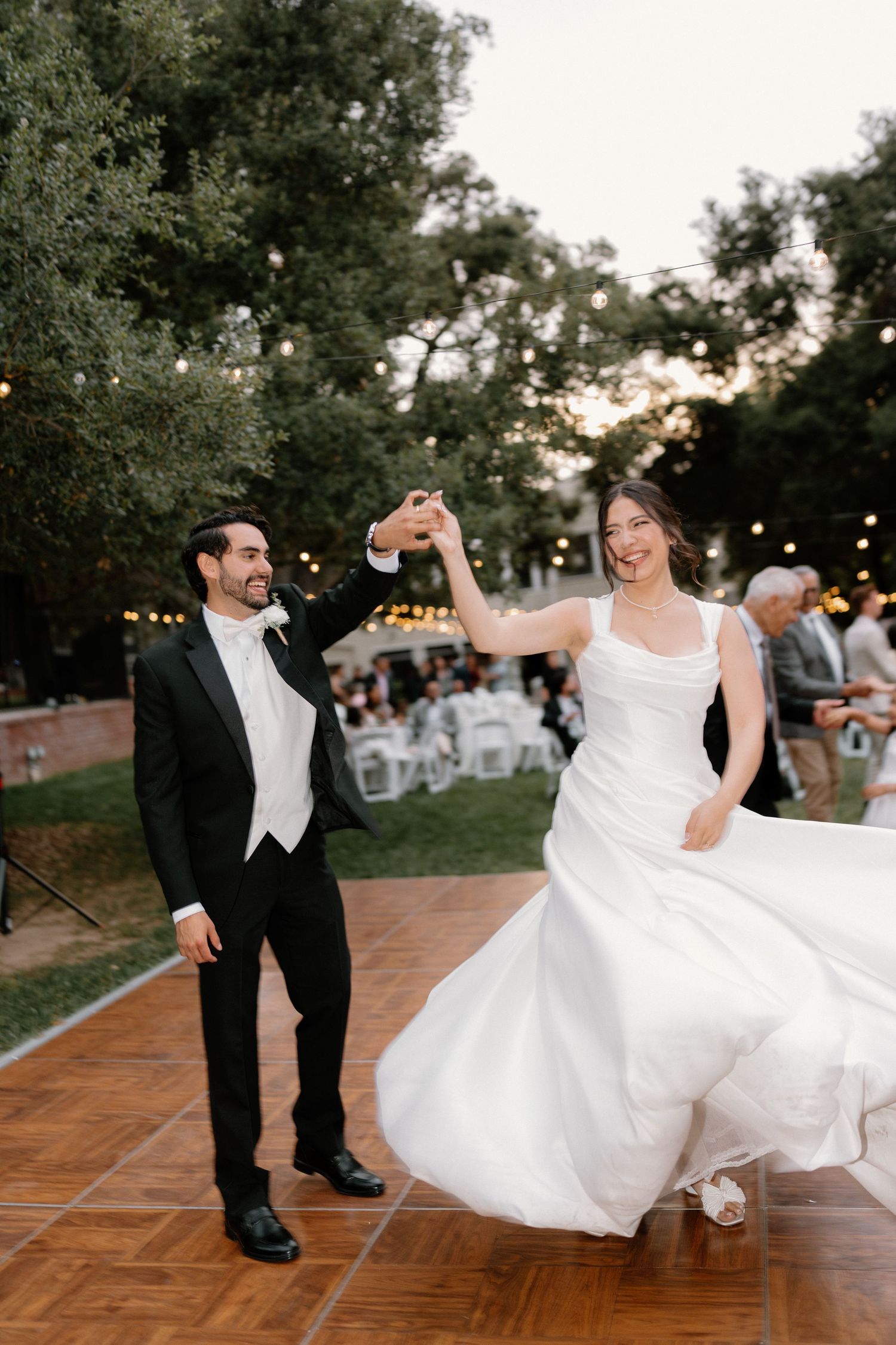 Joyful bride and groom share first dance outdoors at sunset wedding reception with string lights.