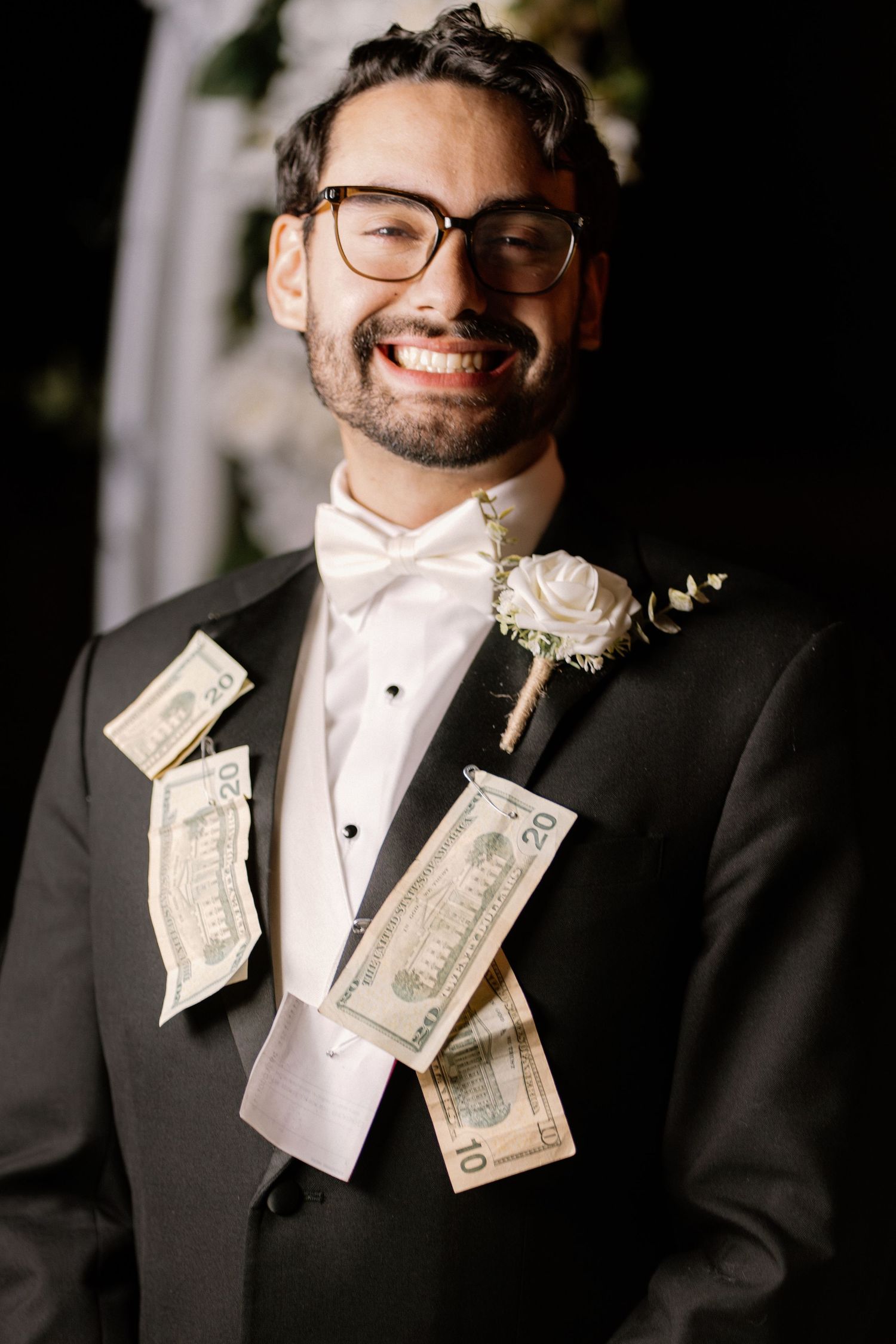Smiling groom in black tuxedo with dollar bills tucked in jacket and white rose boutonniere.