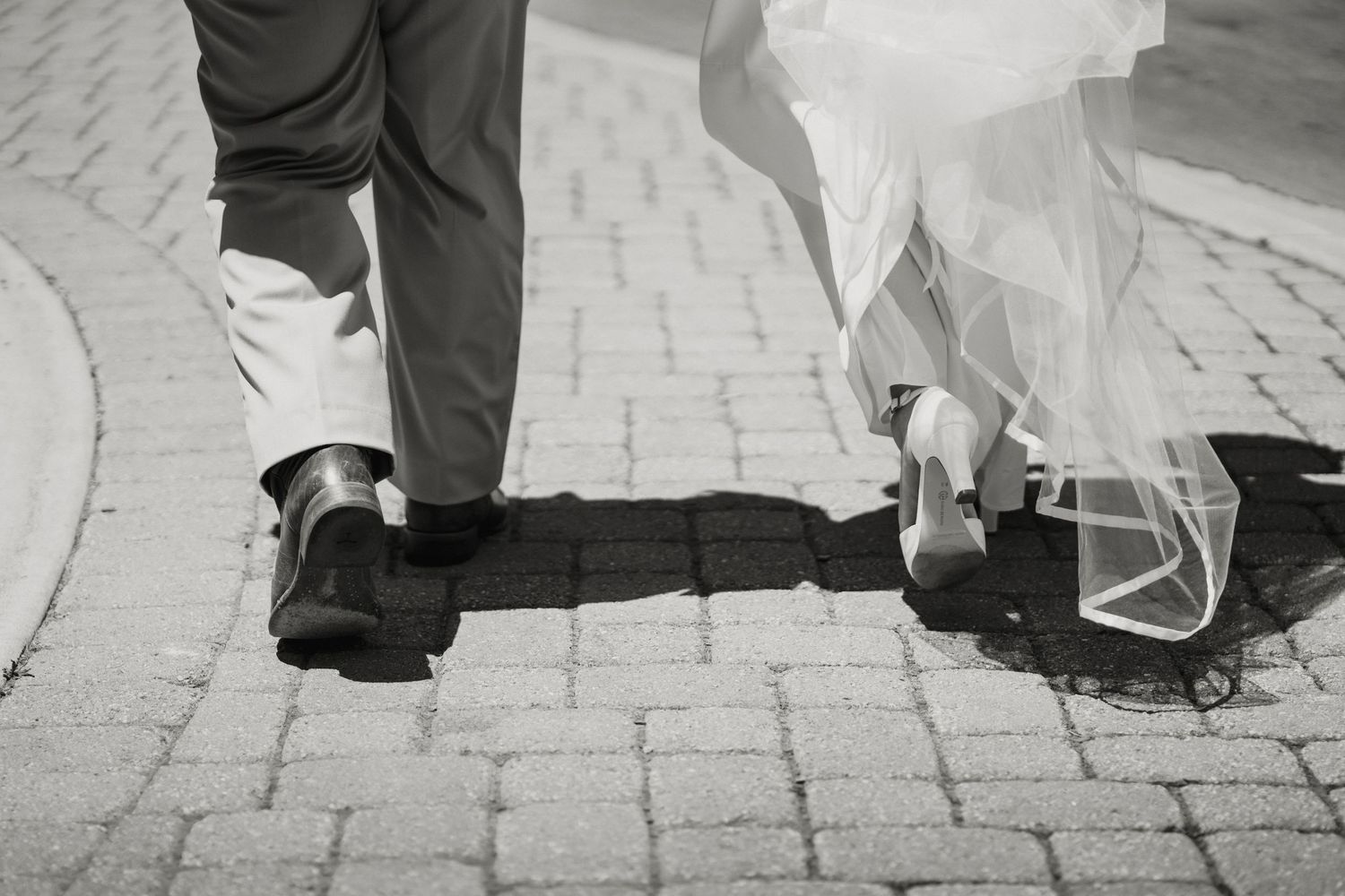 Black and white photo of bride and groom walking on cobblestone, showing wedding dress hem and dress shoes.