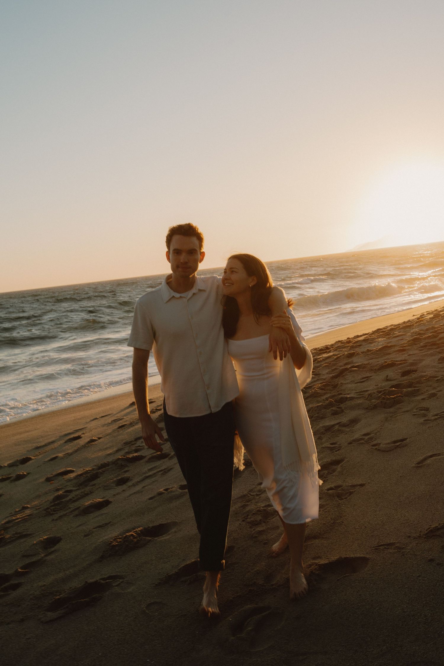 Couple walking barefoot on sandy beach at golden sunset, woman leaning on mans shoulder.