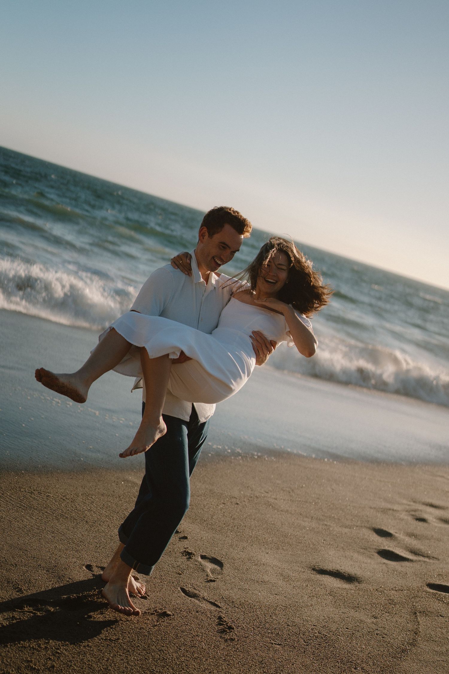 Groom carries laughing bride in his arms on a sandy beach at sunset with ocean waves behind them.