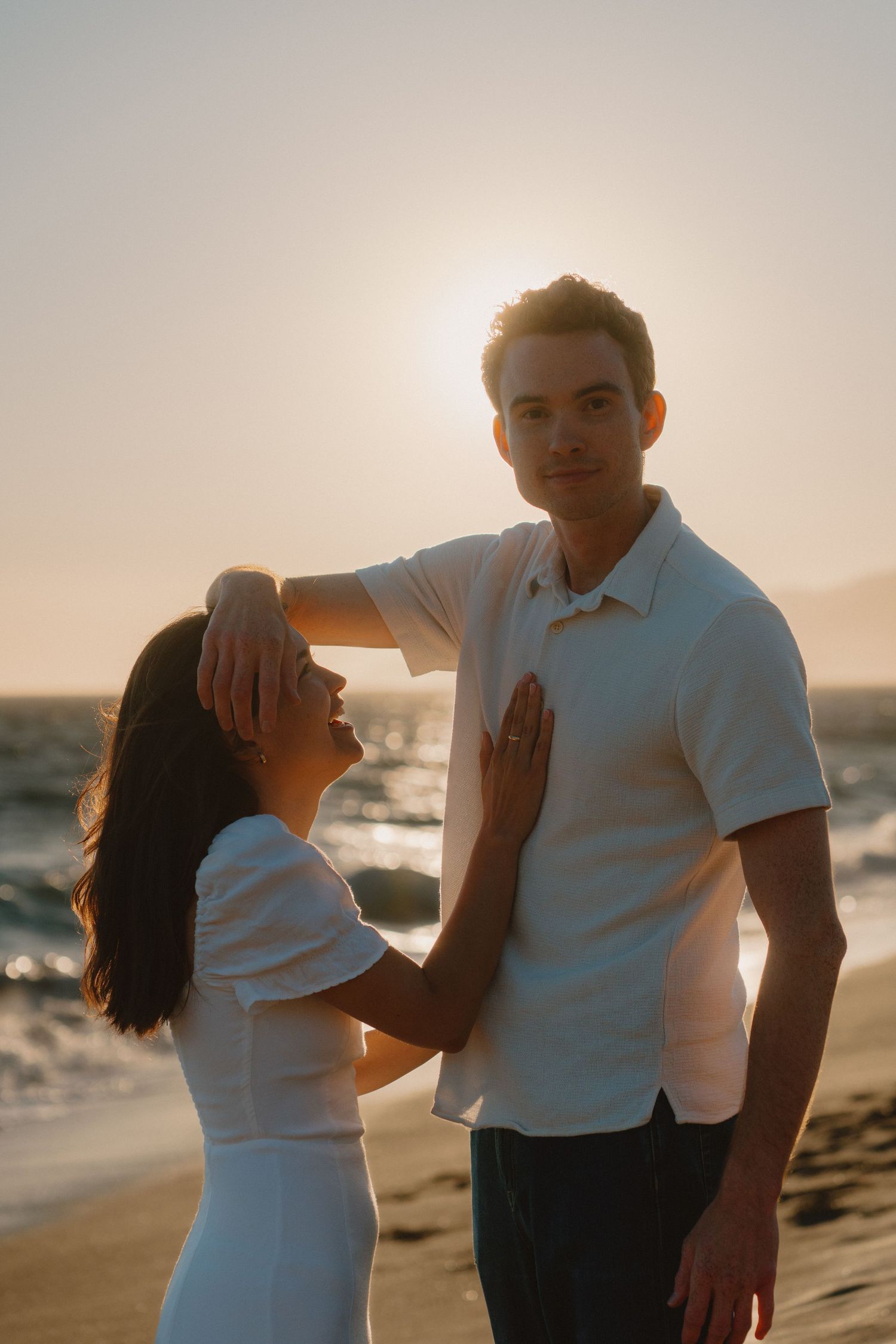 Couple sharing a tender moment on a sunlit beach at golden hour, woman in white dress placing hand on mans chest.