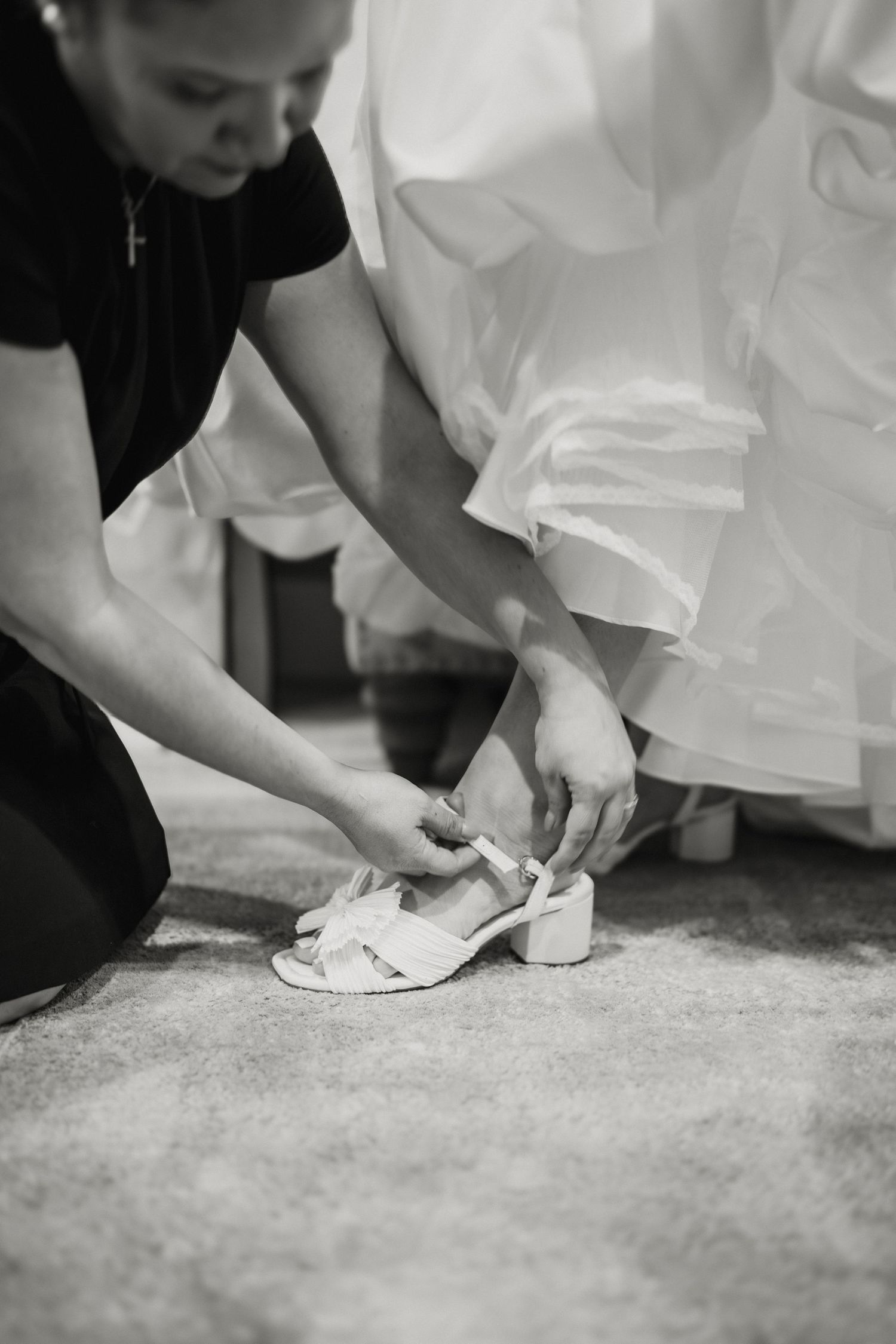 Black and white photo of bridesmaid helping bride fasten white heeled sandals before wedding.