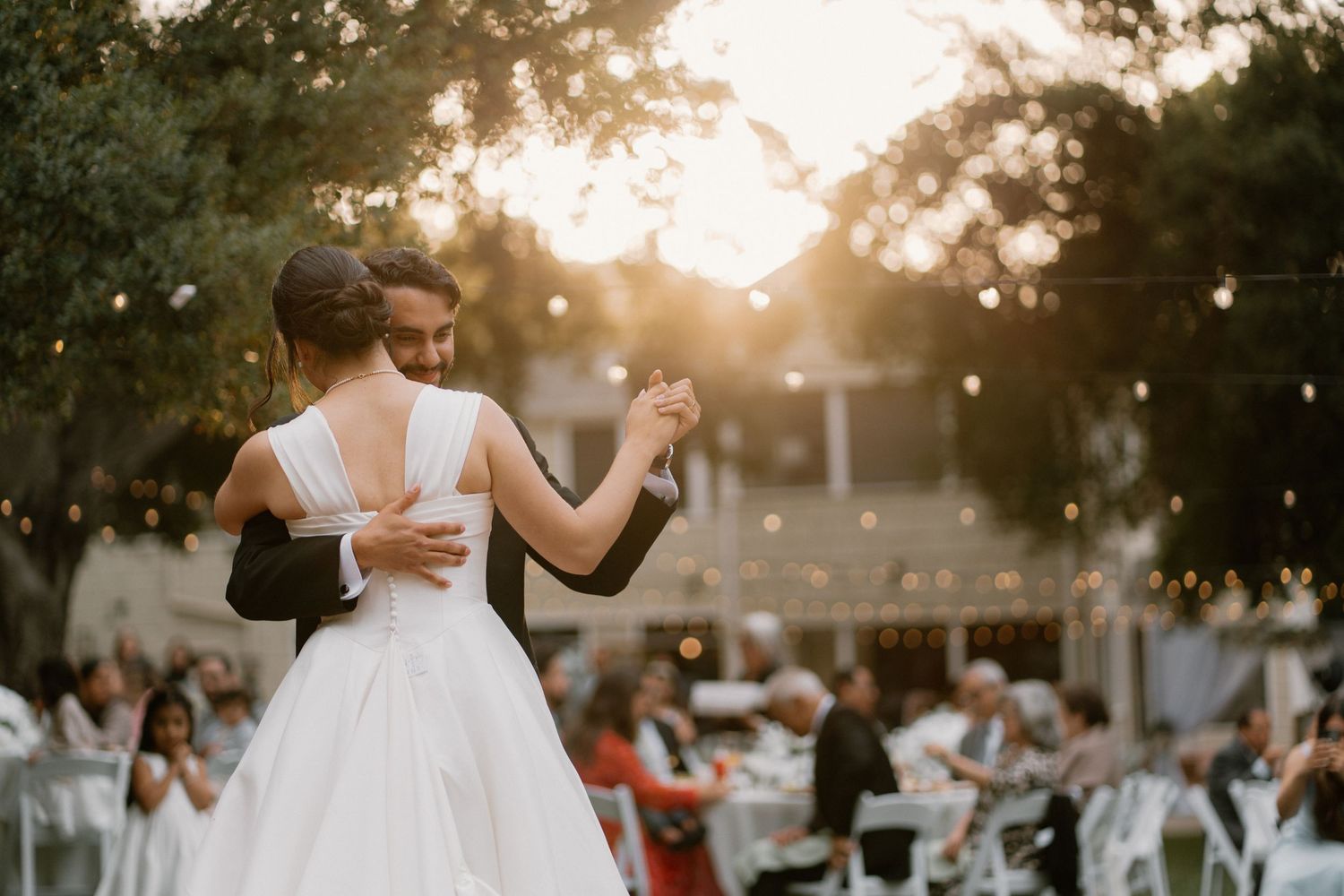 Bride and groom dance outdoors at sunset with string lights and wedding guests seated in background.