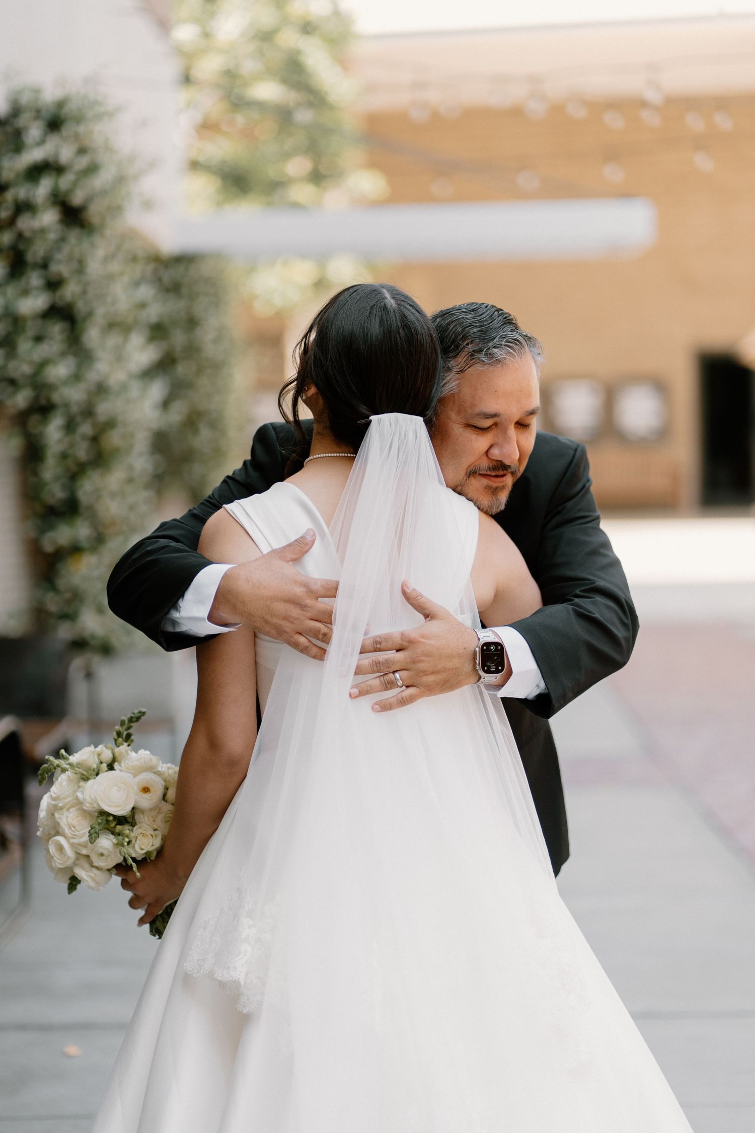 Emotional father embraces bride in white wedding gown and veil during heartfelt first look moment.