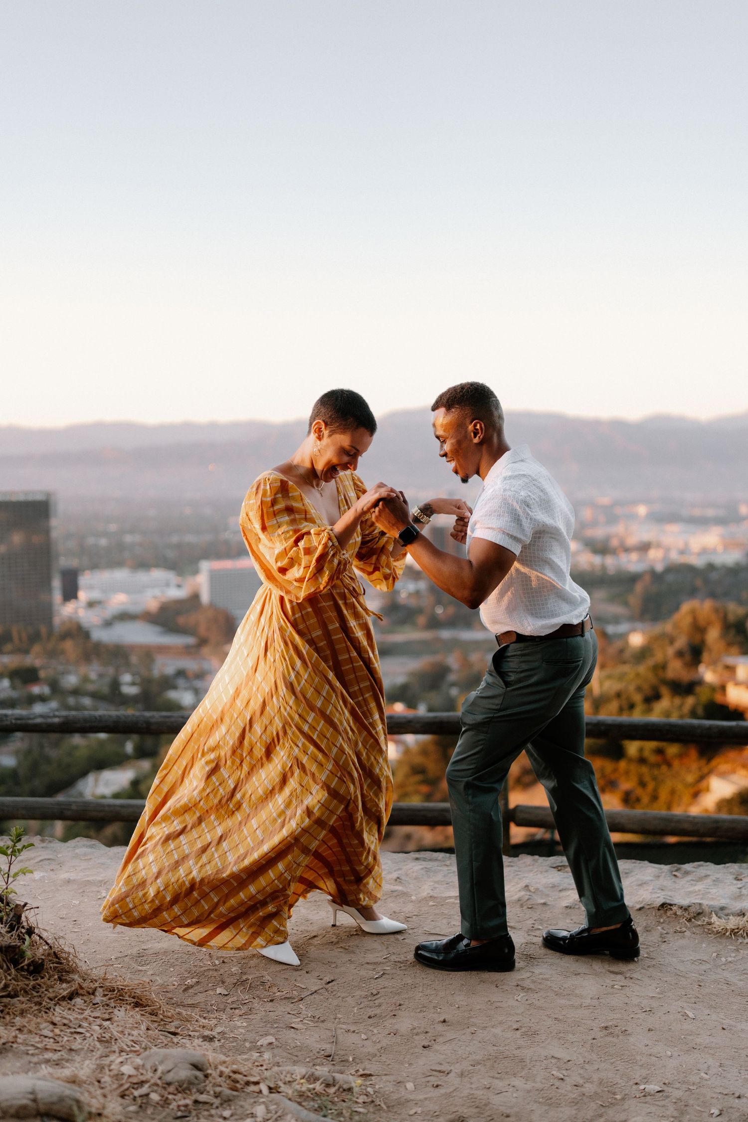 Couple dancing at golden hour on a hilltop overlook with city and mountain views, woman in flowing gold dress.
