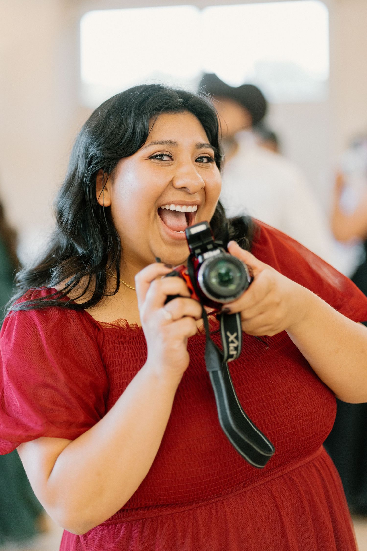 Happy woman in red dress laughing while holding DSLR camera at wedding celebration.