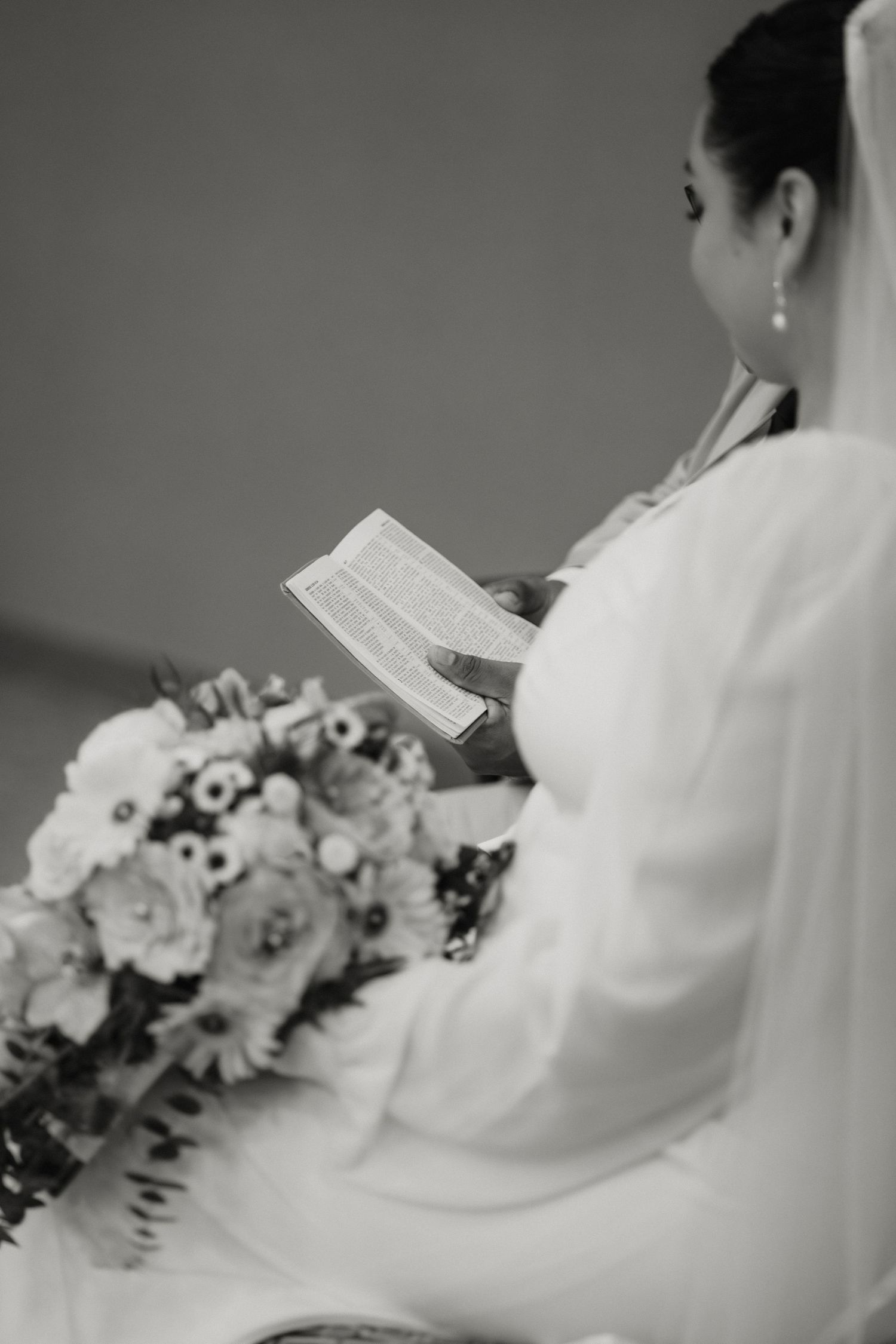 Black and white photo of bride in veil reading from book while holding large floral bouquet at wedding ceremony.