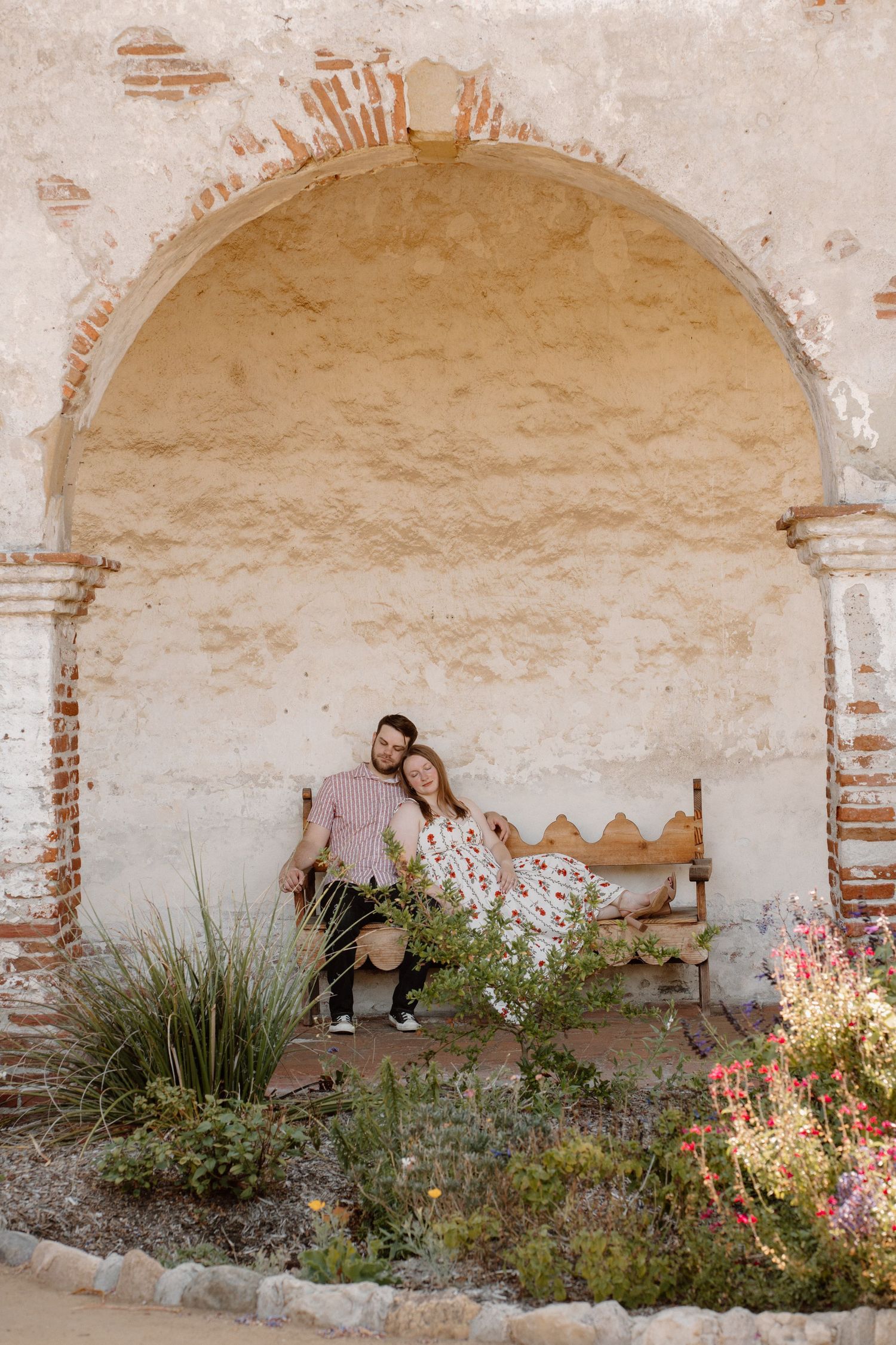 Couple sitting together on a bench inside a rustic brick archway surrounded by colorful garden flowers.