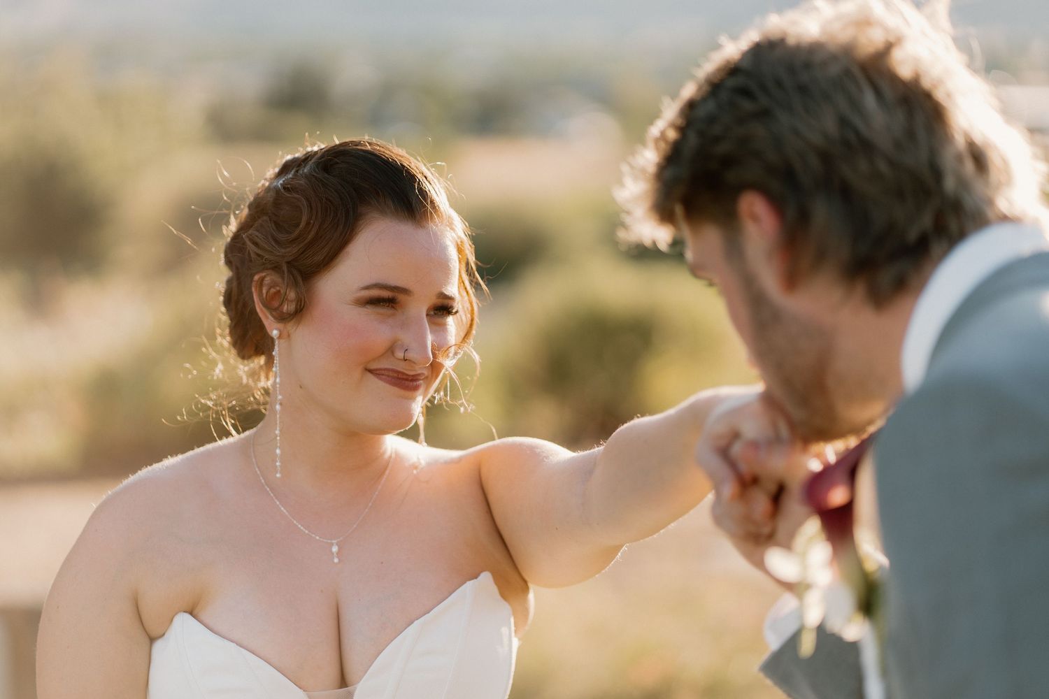 Bride smiling as groom kisses her hand during outdoor wedding ceremony in warm sunlight.