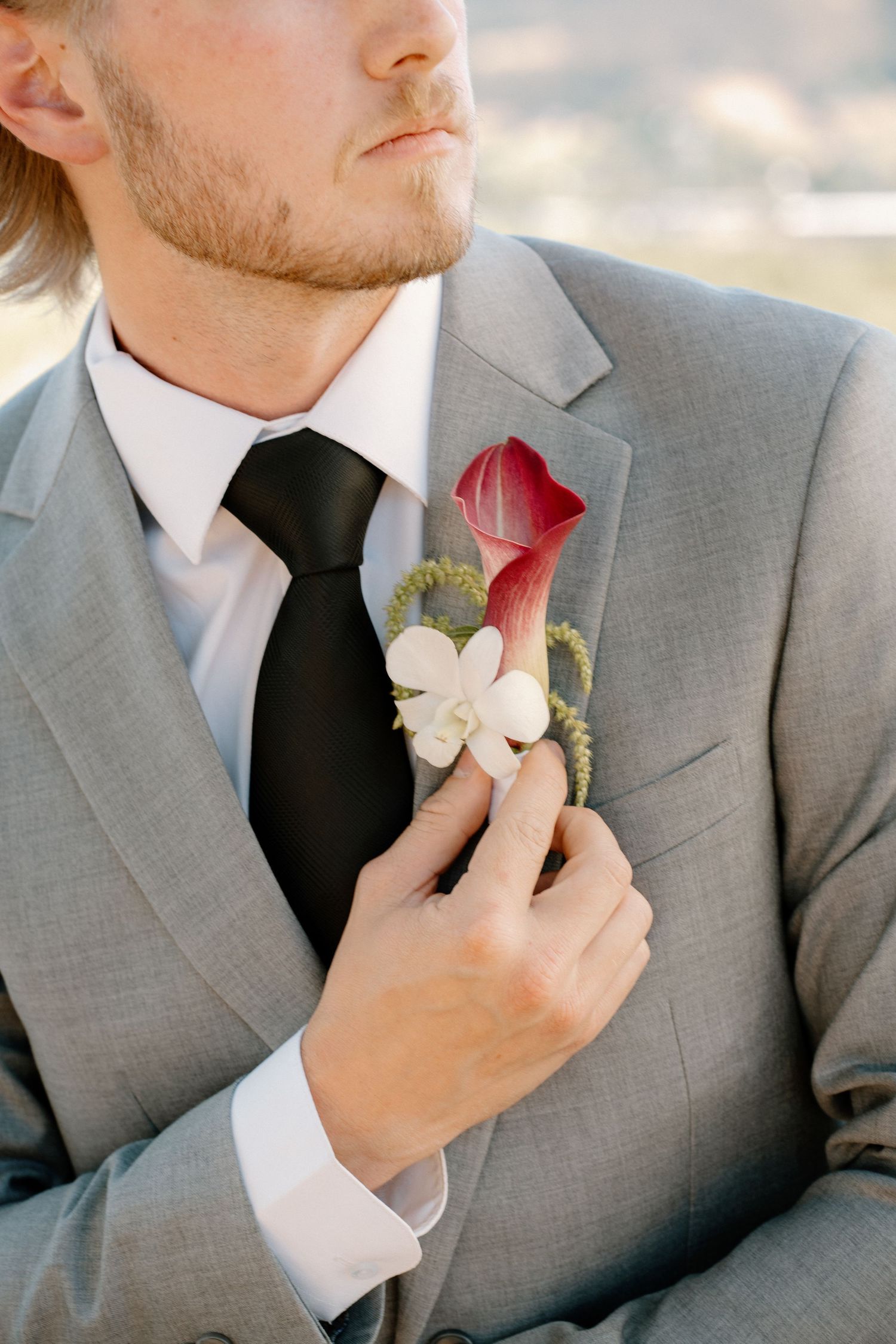 Groom in gray suit adjusting red calla lily and white orchid boutonniere on lapel outdoors.