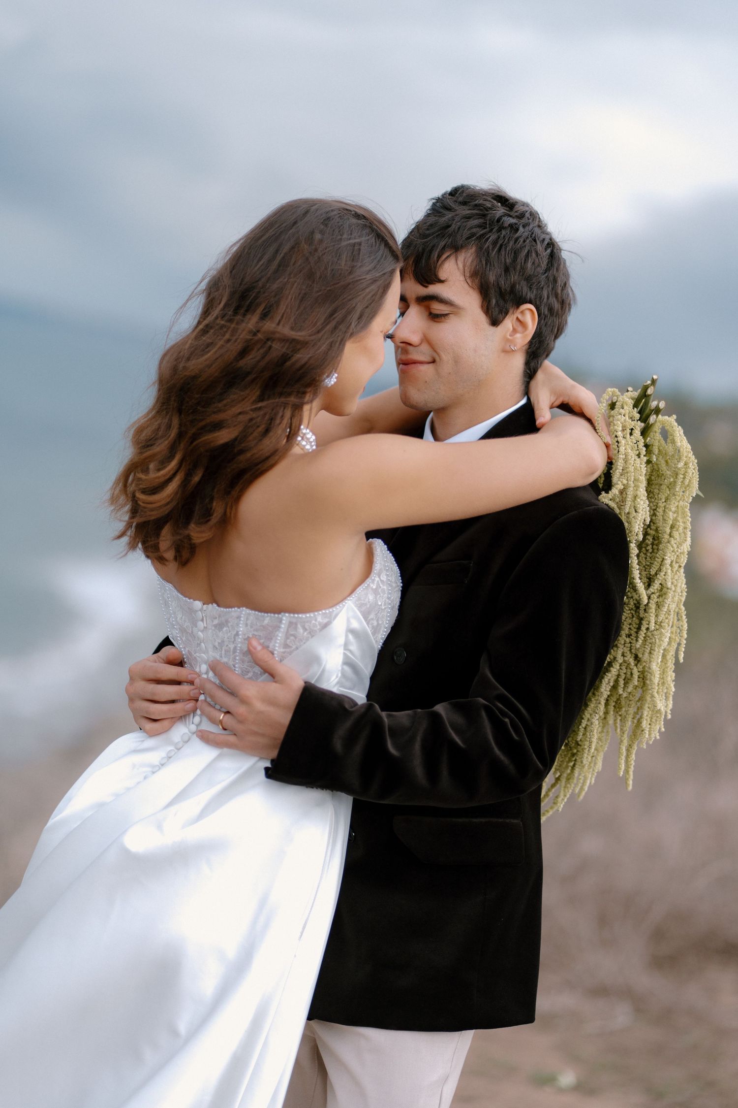 Groom embraces bride in white strapless gown near scenic ocean shoreline.