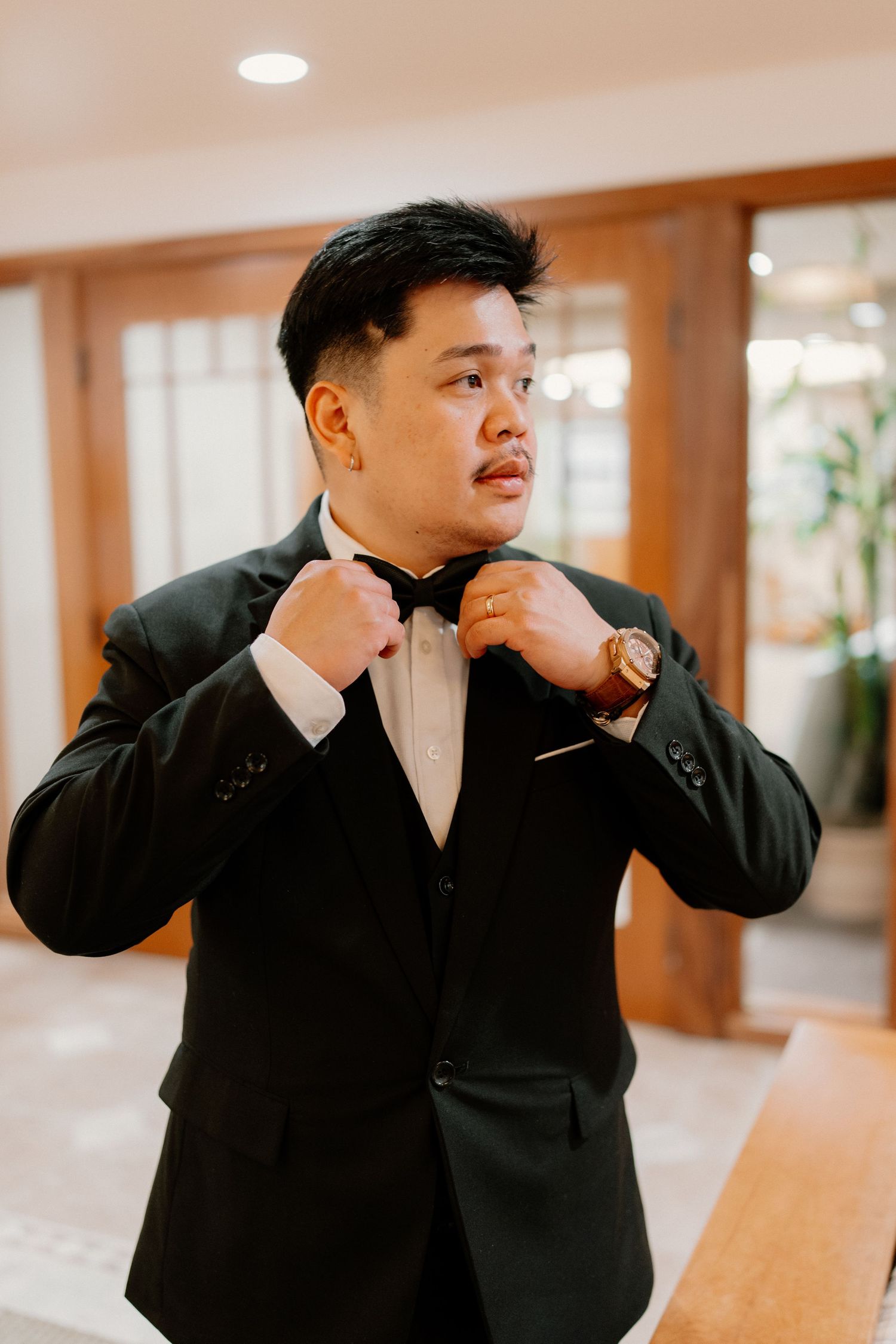 Dapper groom in a black tuxedo adjusting his bow tie while getting ready for his wedding.