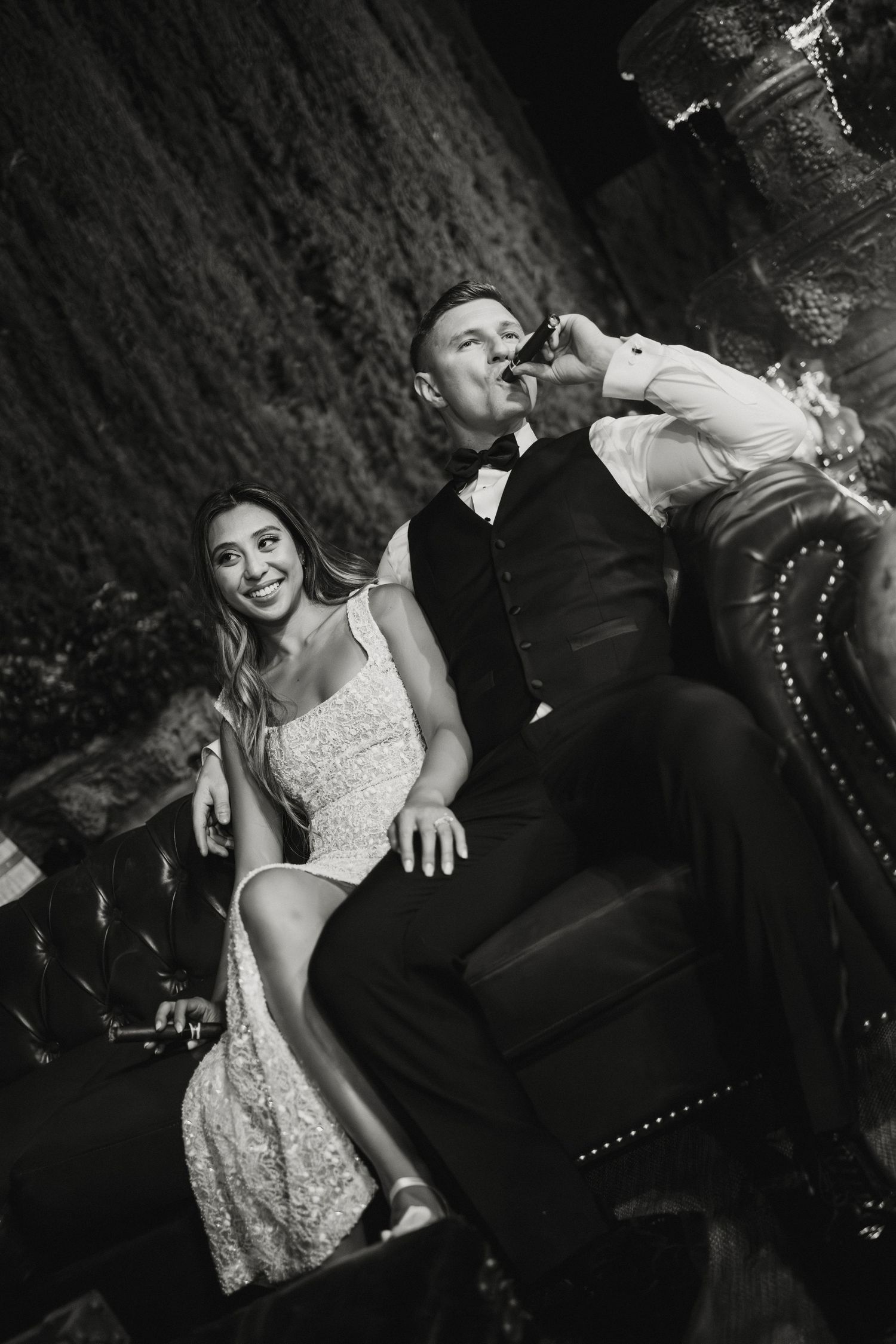 Black and white photo of bride and groom relaxing on leather sofa, groom drinking a toast at wedding reception.