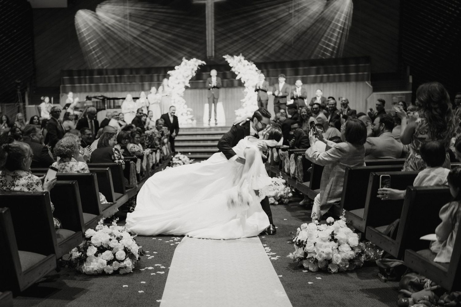 Black and white photo of newlyweds kissing at altar with floral arch in church ceremony.