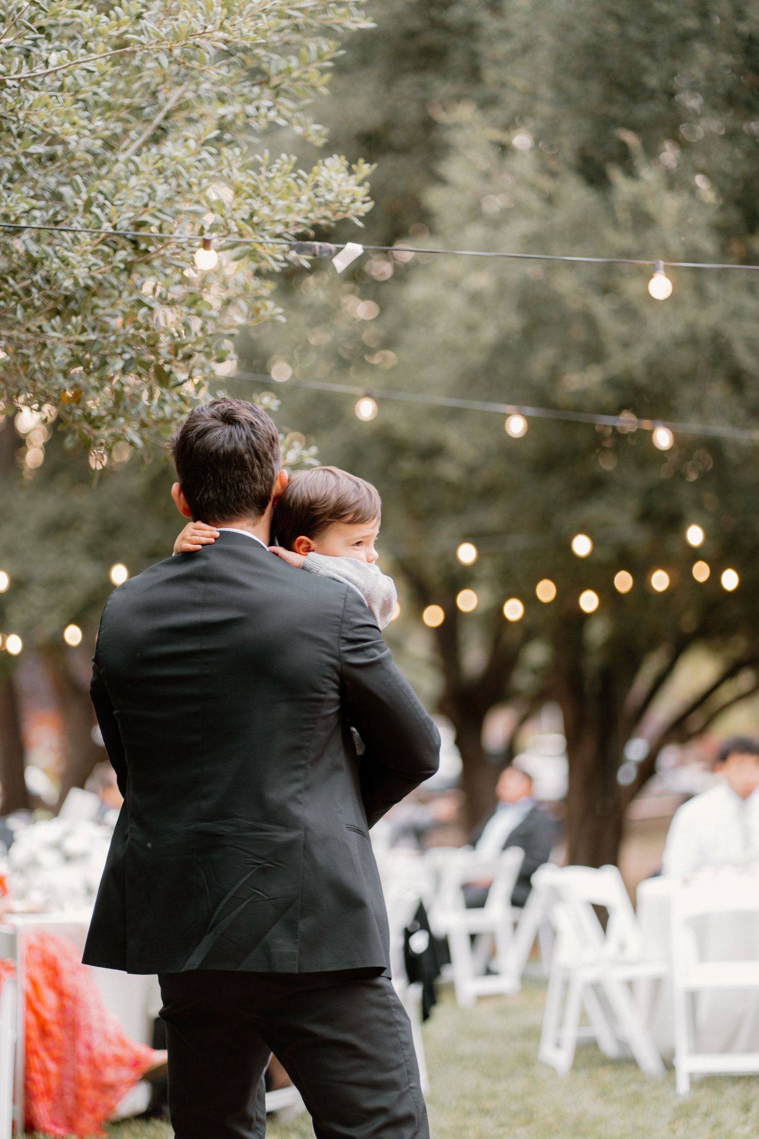 Groom in black suit holds young child at outdoor wedding reception with string lights.
