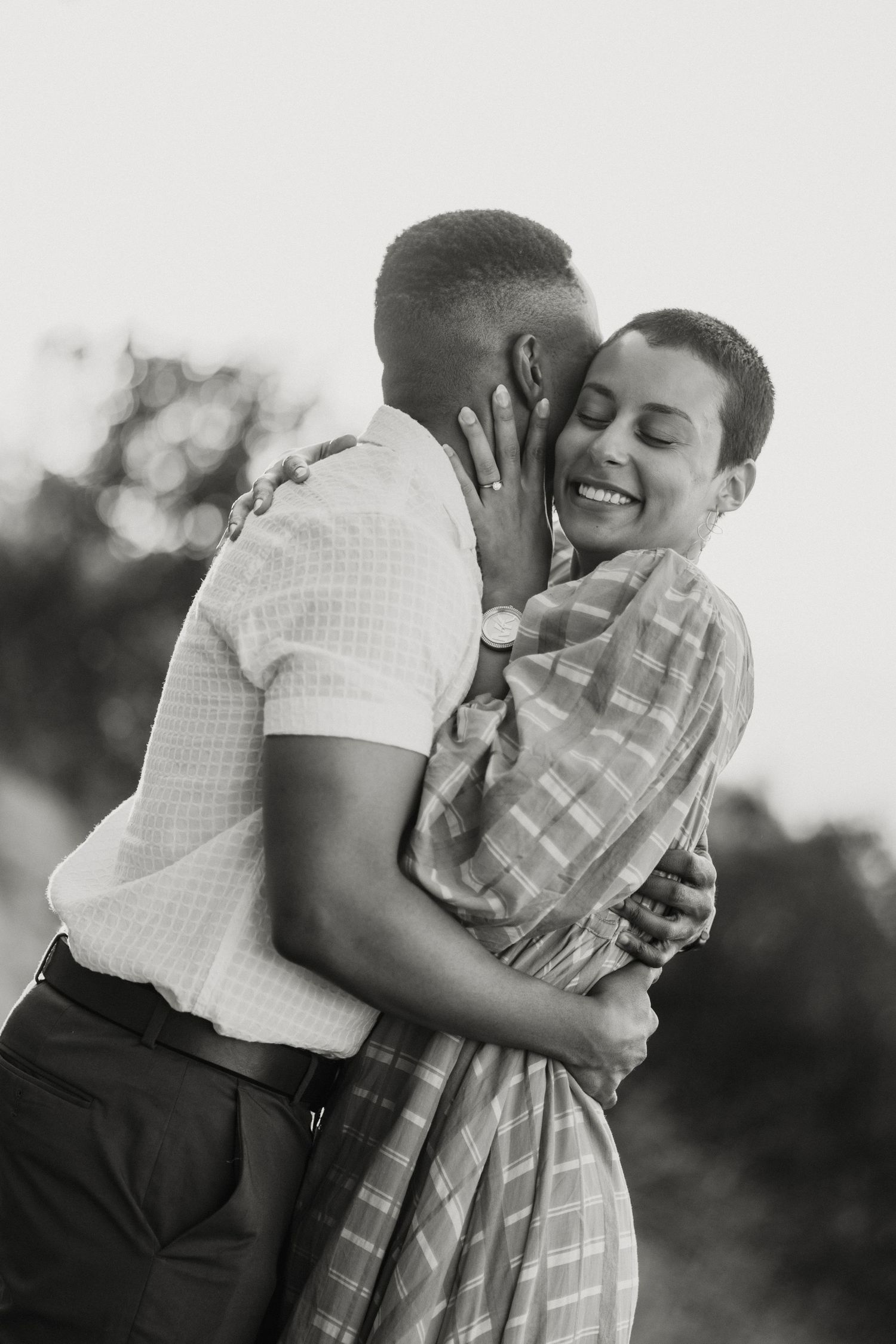 Black and white photo of a joyful couple embracing outdoors, woman smiling as man kisses her cheek.