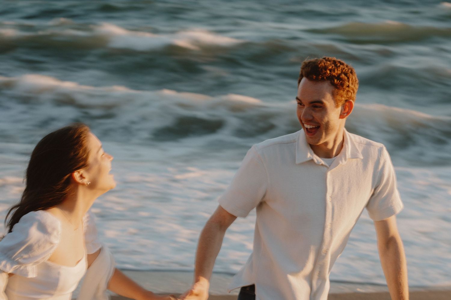 Couple laughing and holding hands on a sunny beach with ocean waves in the background.