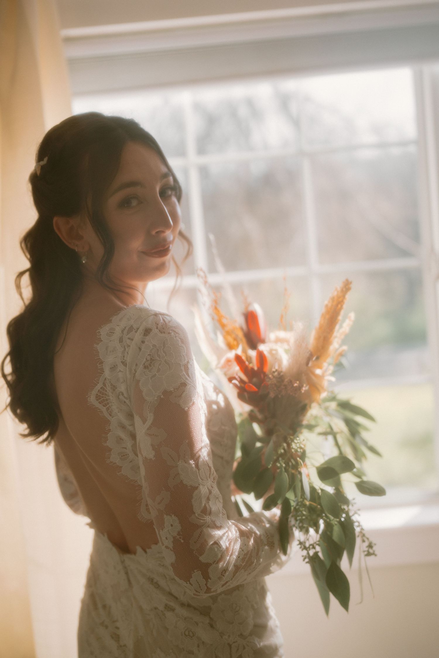 Bride in backless lace wedding dress glancing over shoulder holding autumn bouquet by bright window.
