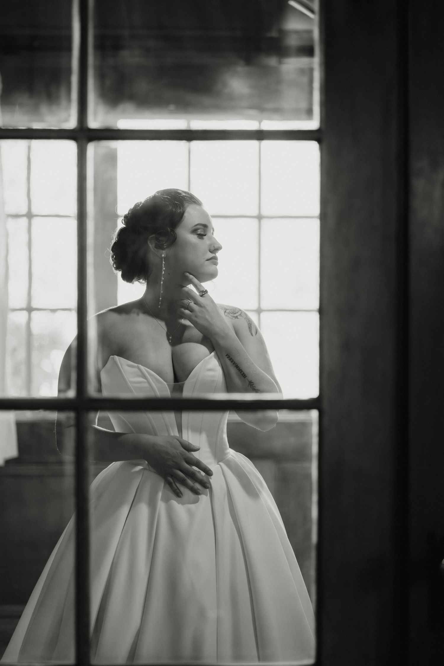 Black and white photo of an elegant bride in a strapless ball gown, gazing thoughtfully beside a large window.