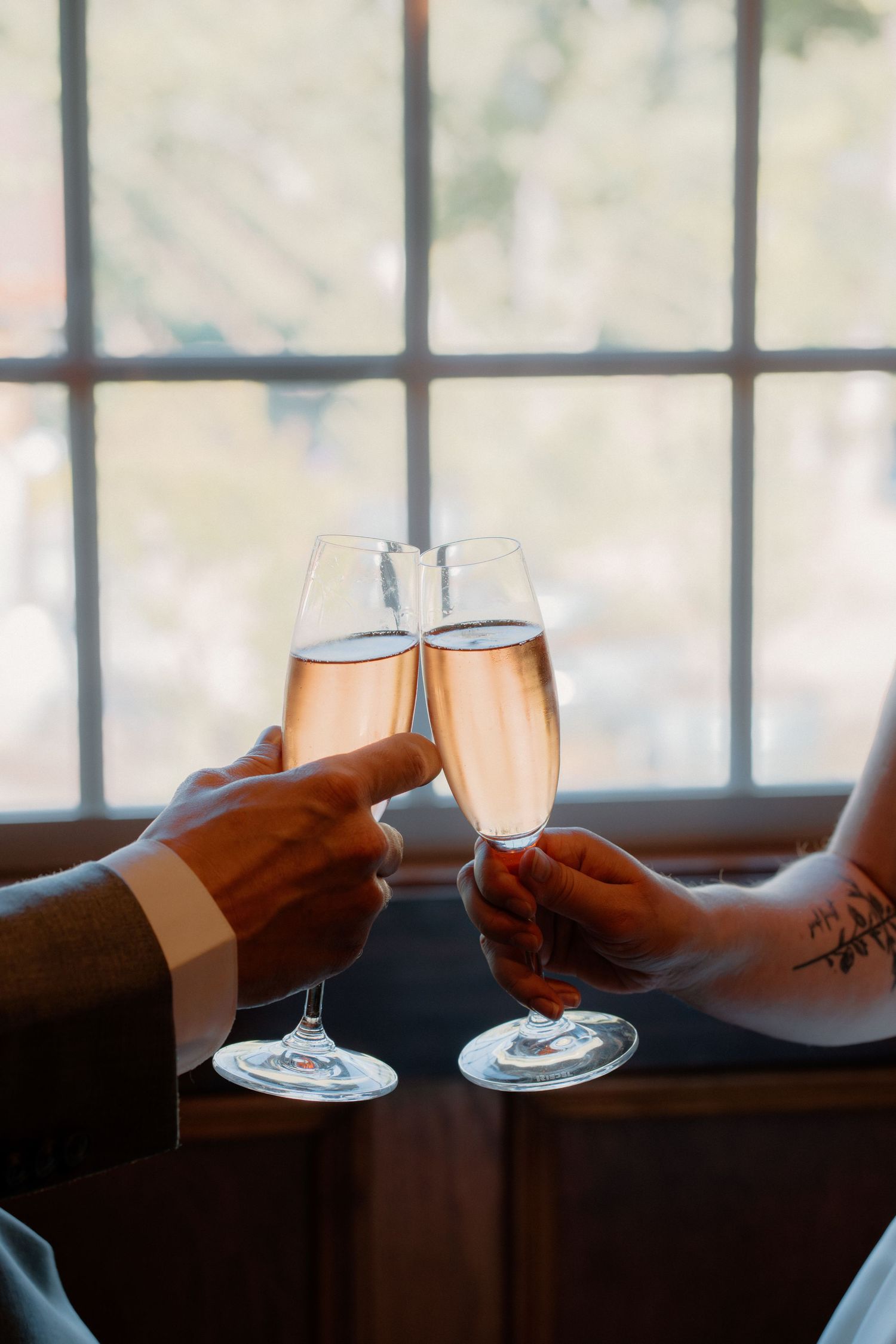 Bride and groom clinking champagne glasses in toast by sunlit window on wedding day.