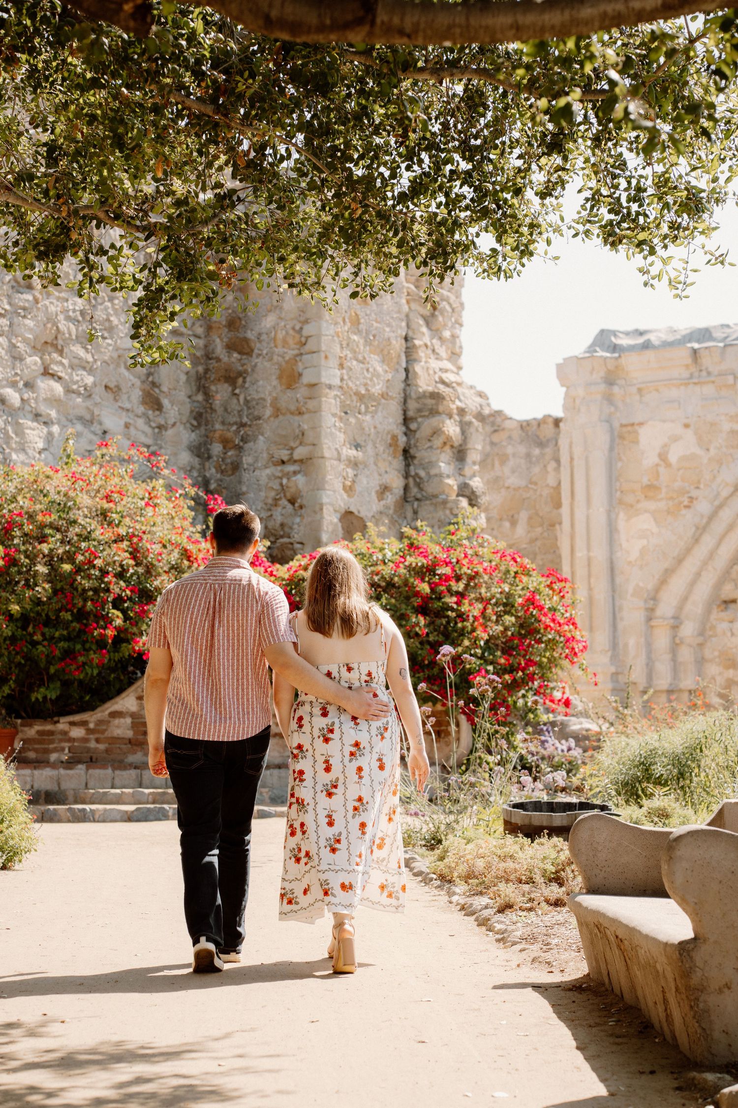 Couple walking hand in hand through a sunlit mission garden with vibrant red bougainvillea blooms.