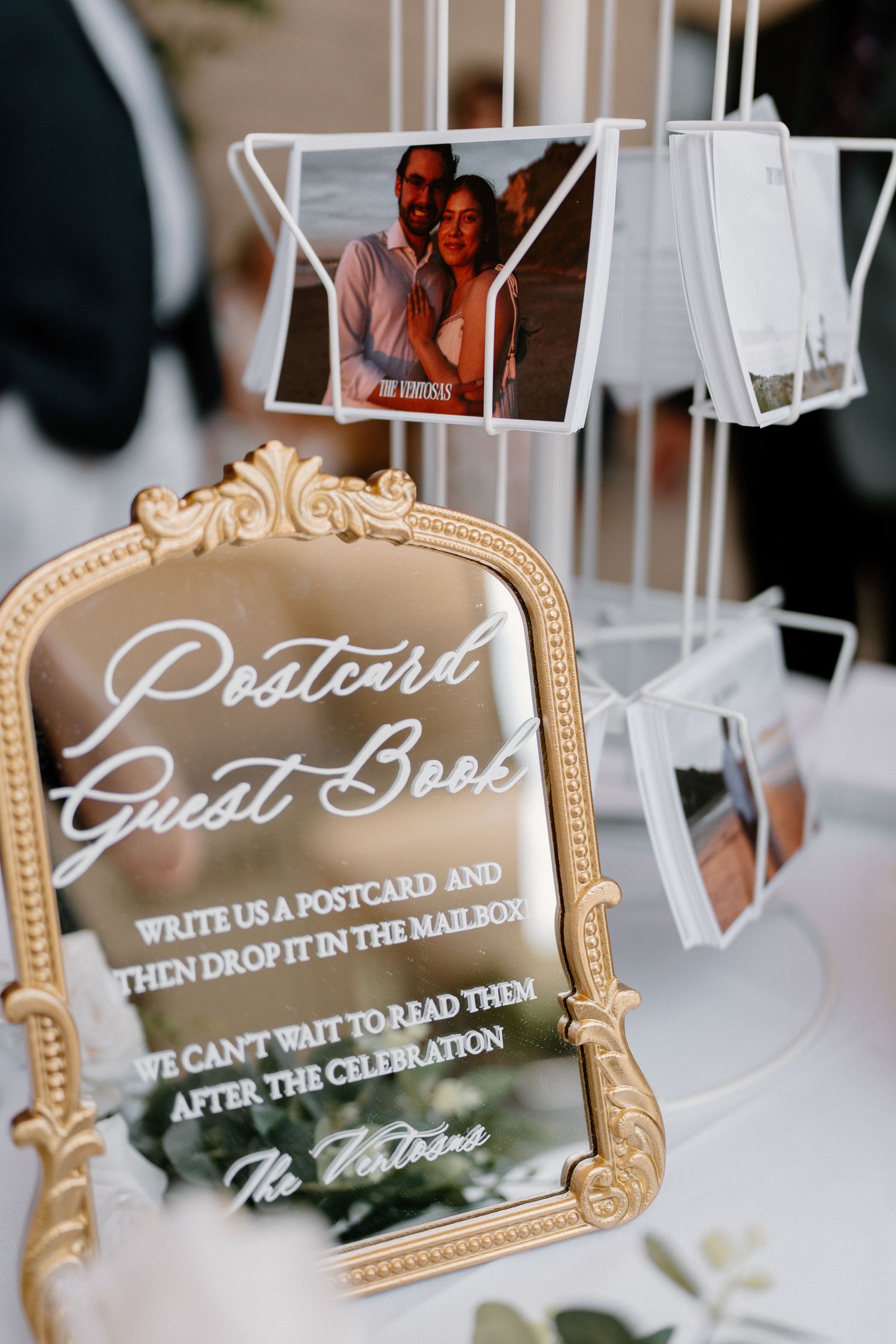 Gold ornate mirror postcard guest book sign with polaroid photos displayed on a white wire stand.